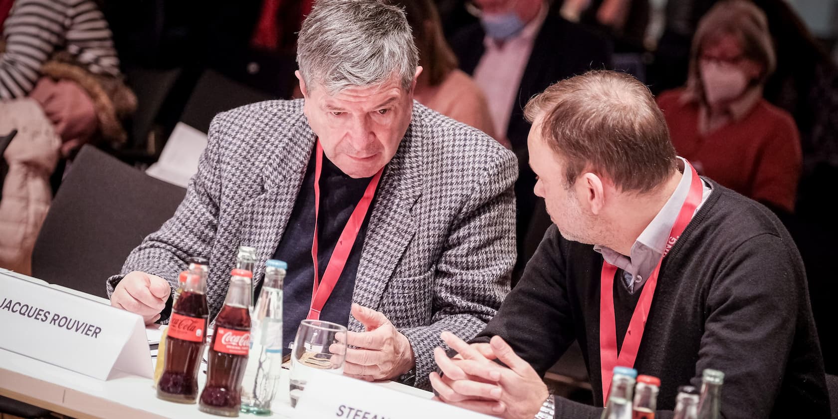 Two men are sitting at a conference table having a conversation. In the foreground, there are several drink bottles and name tags, including one with the name 'Jacques Rouvier'.