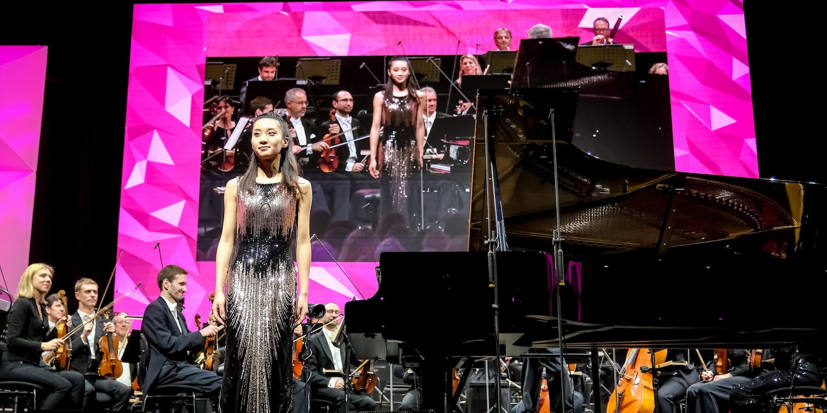 A musician stands in the foreground in front of a grand piano, surrounded by an orchestra. In the background, a large screen displays her image.