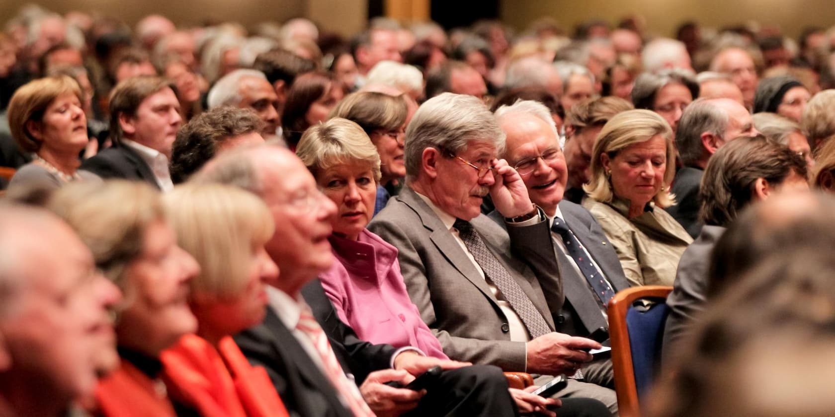 Audience in a crowded event hall, an elderly person in the middle adjusting their glasses.