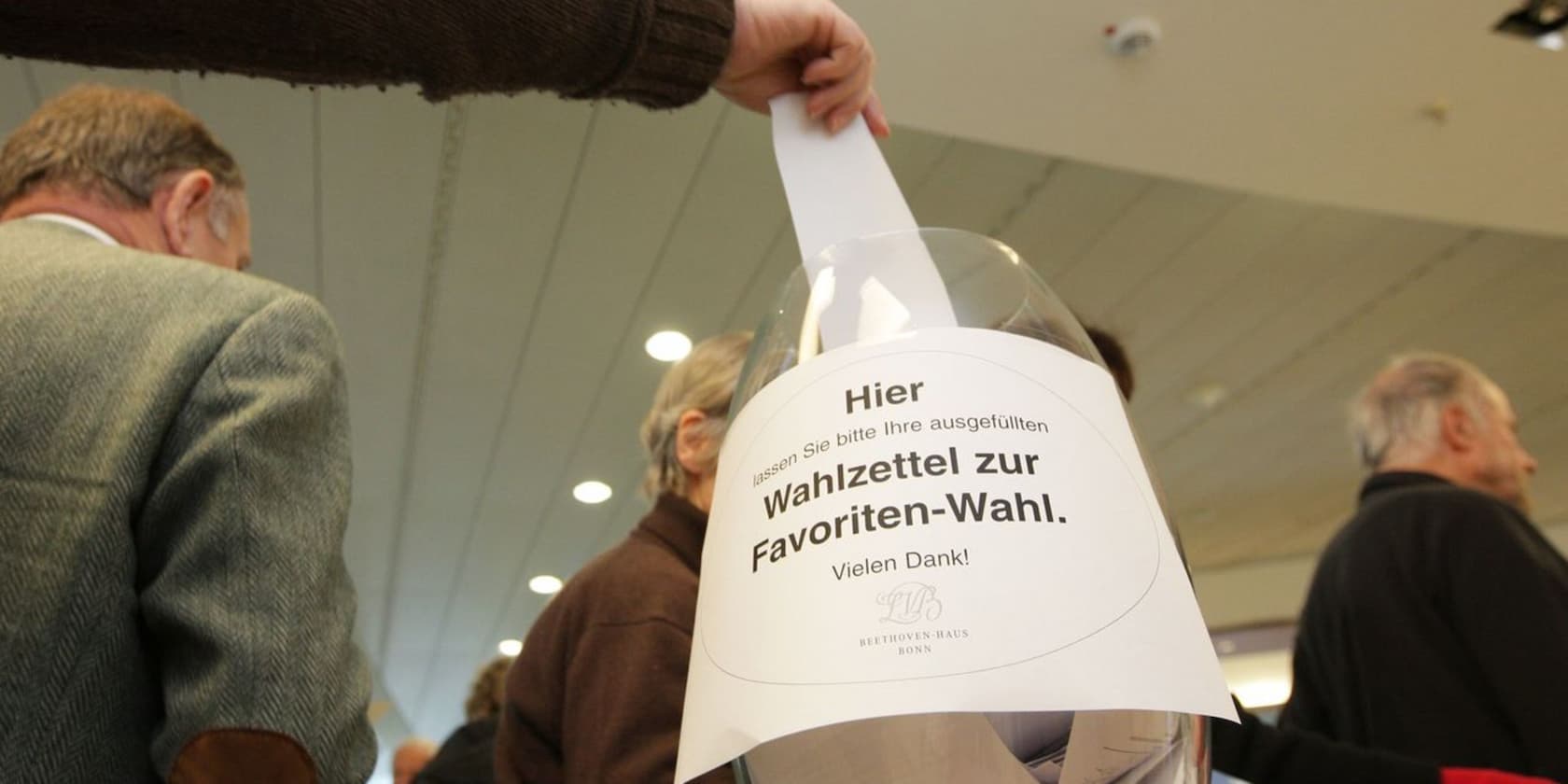 A person places a completed ballot into a ballot box for the favorites vote. Several people stand in the background.