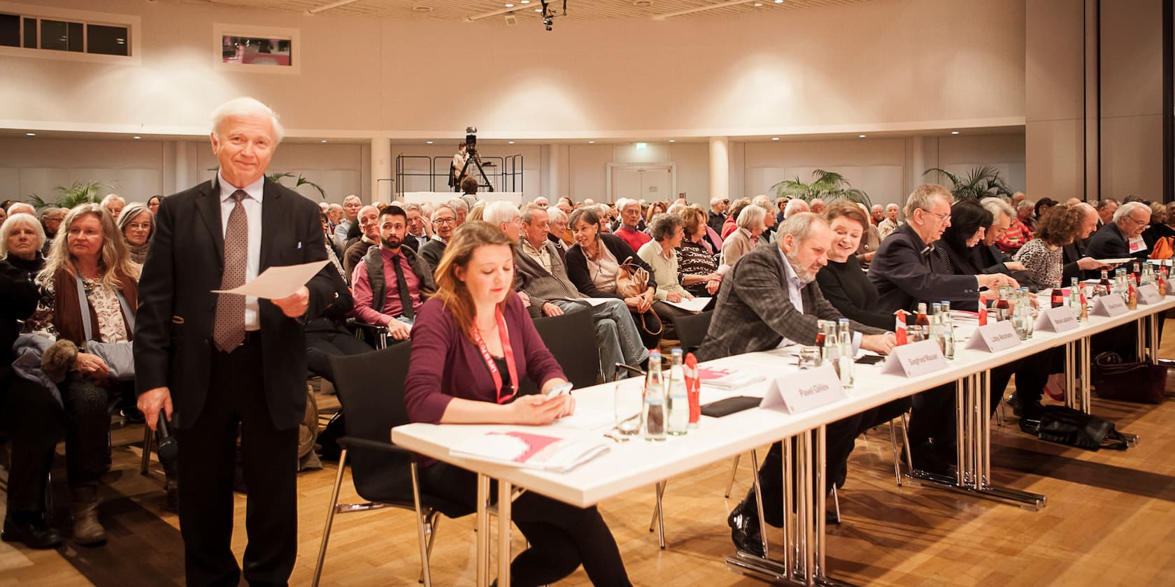 People at an event in a conference room, a man stands in the foreground holding a piece of paper