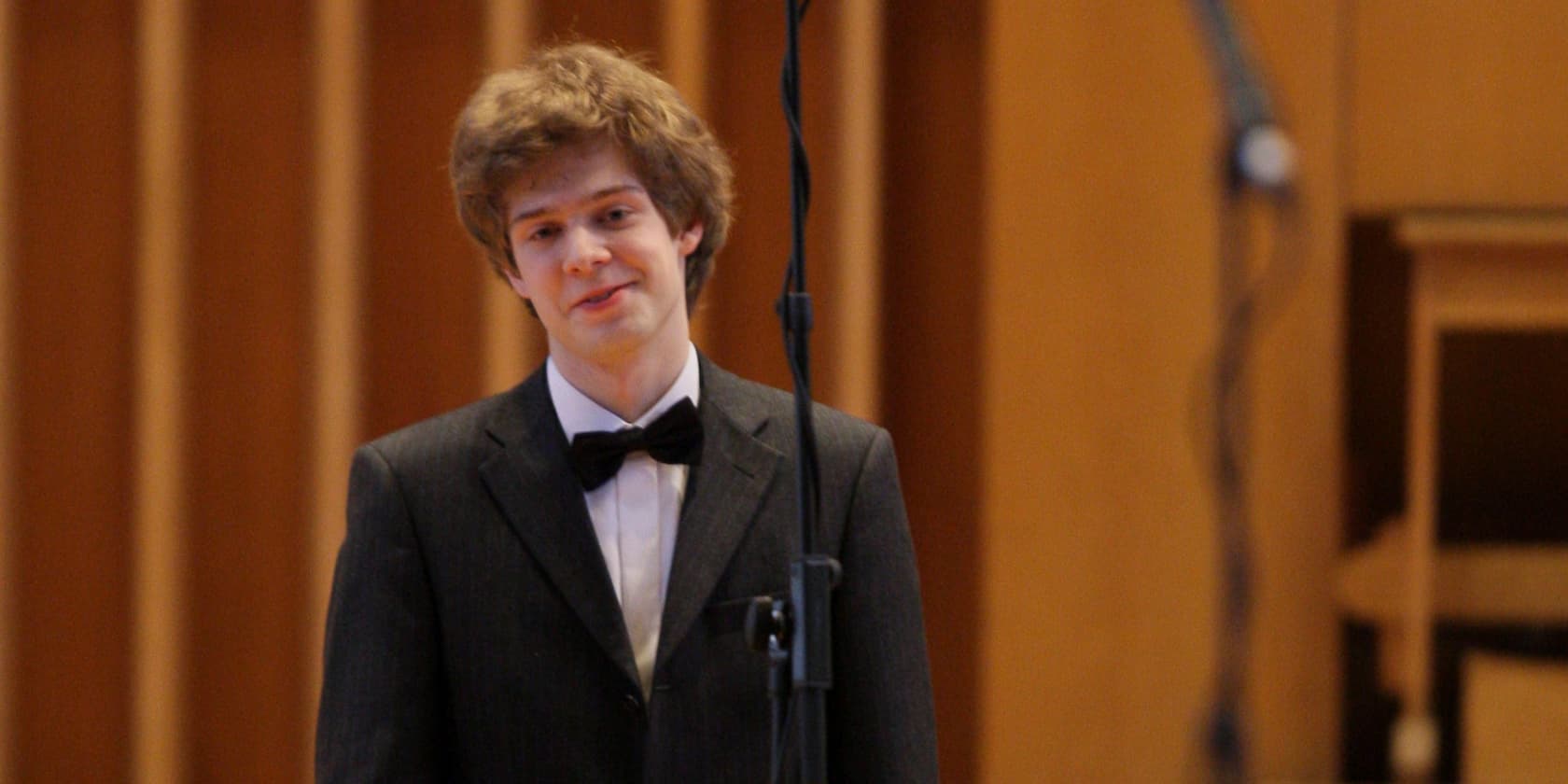 A young man in a suit with a bow tie stands in front of a microphone.