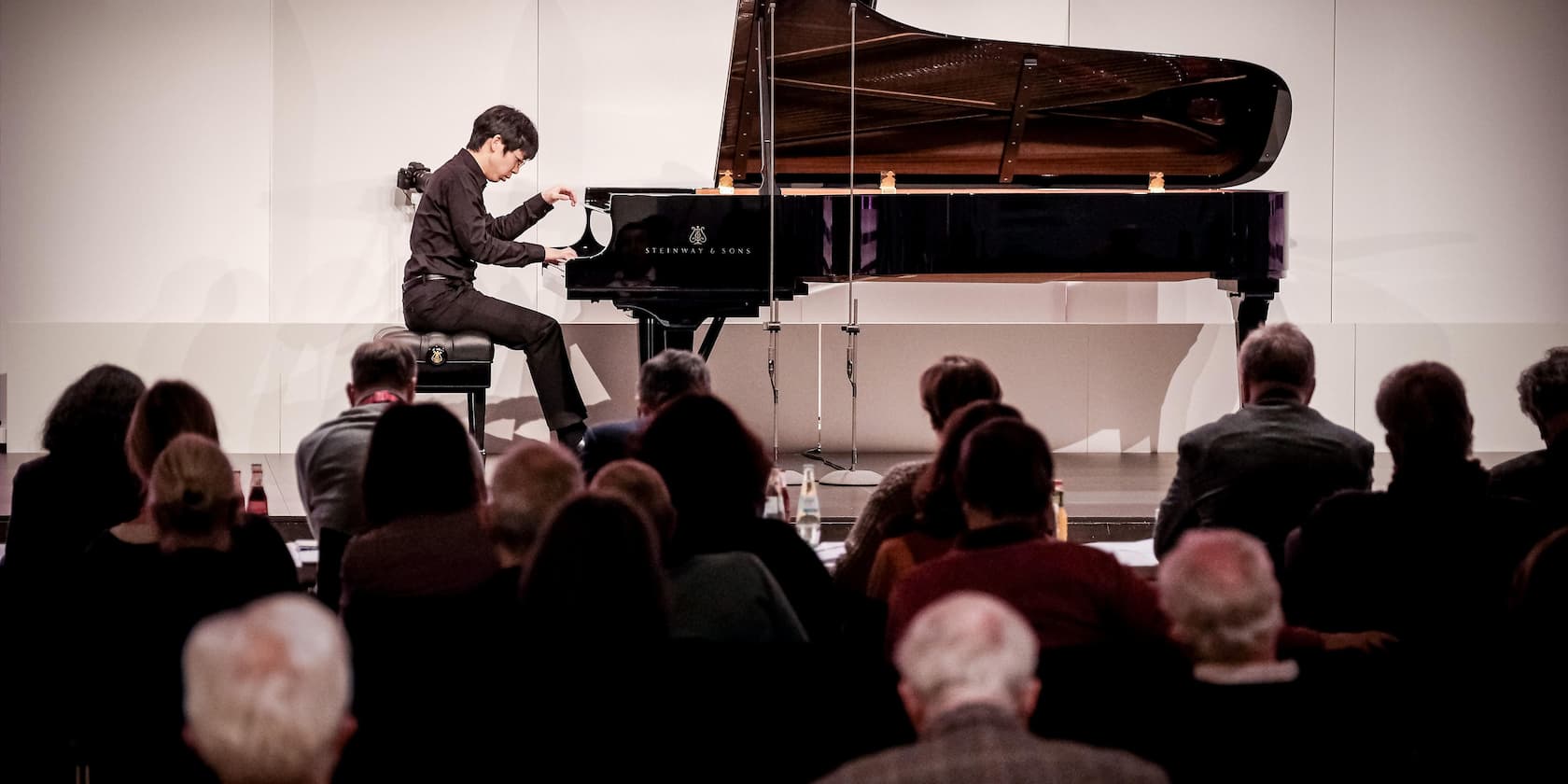 A pianist plays on a grand piano in front of a seated audience.