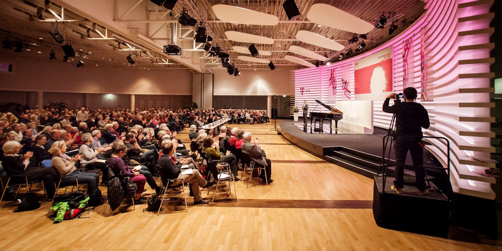 Konferenzsaal mit Publikum, das einem Redner auf der Bühne zuhört und klatscht. Ein Kameramann filmt die Veranstaltung.