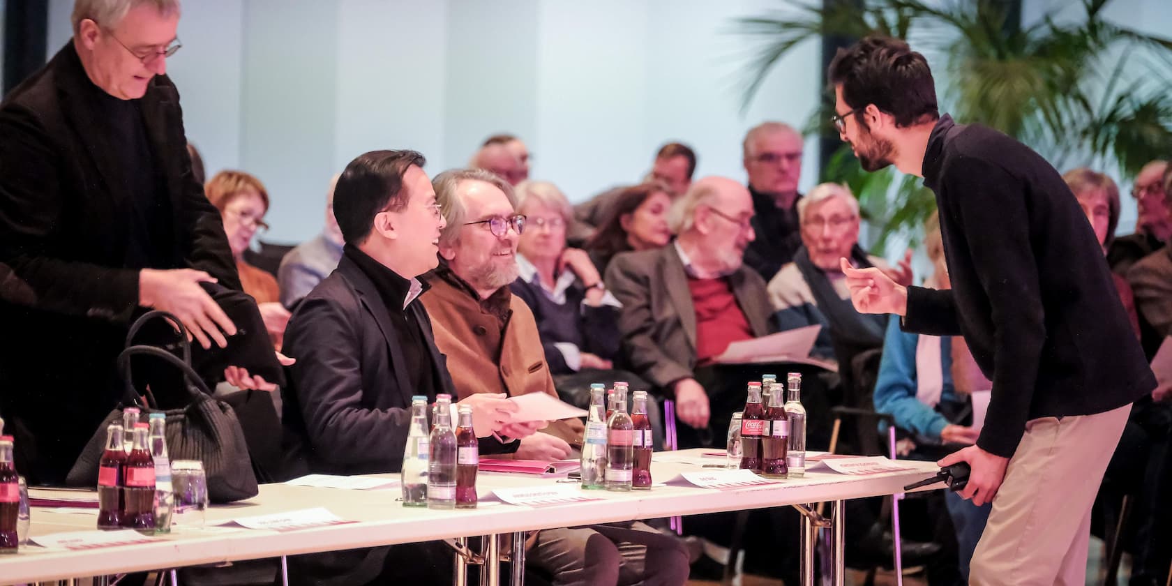 People sitting at a table and talking at an event, with an audience seated in the background.