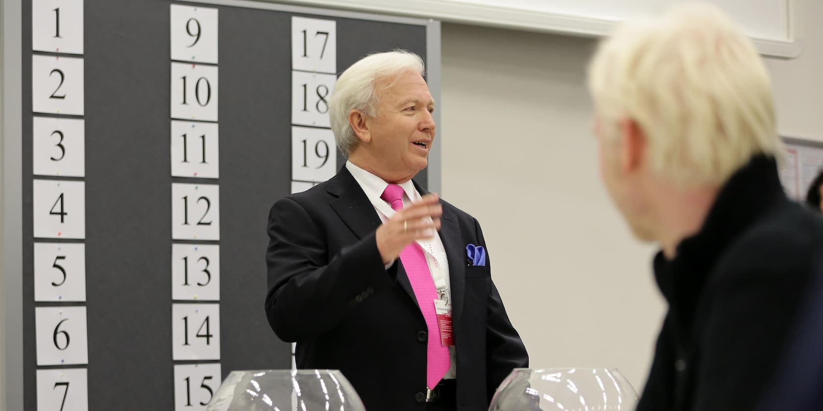 A man in a black suit and pink tie speaks in front of a wall with numbered cards while another person listens in the foreground.