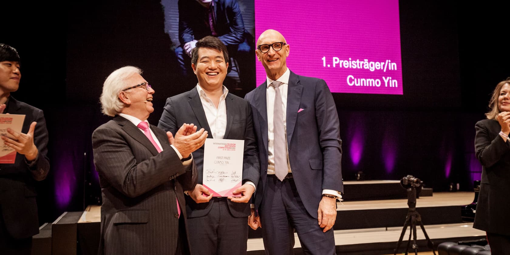 Four people standing on a stage and smiling. The person in the center holds a certificate that reads 'FIRST PRIZE, CUNMO YIN'. In the background, a board reads '1. Preisträger/in Cunmo Yin'.