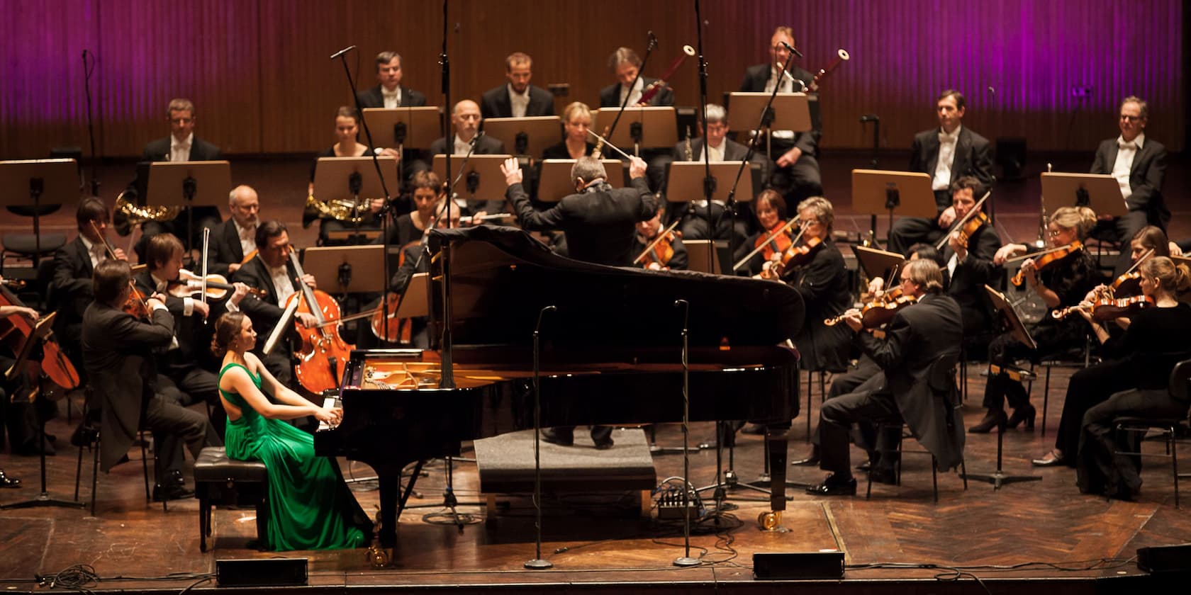 A woman in a green dress plays the piano on stage, accompanied by an orchestra conducted by a conductor.