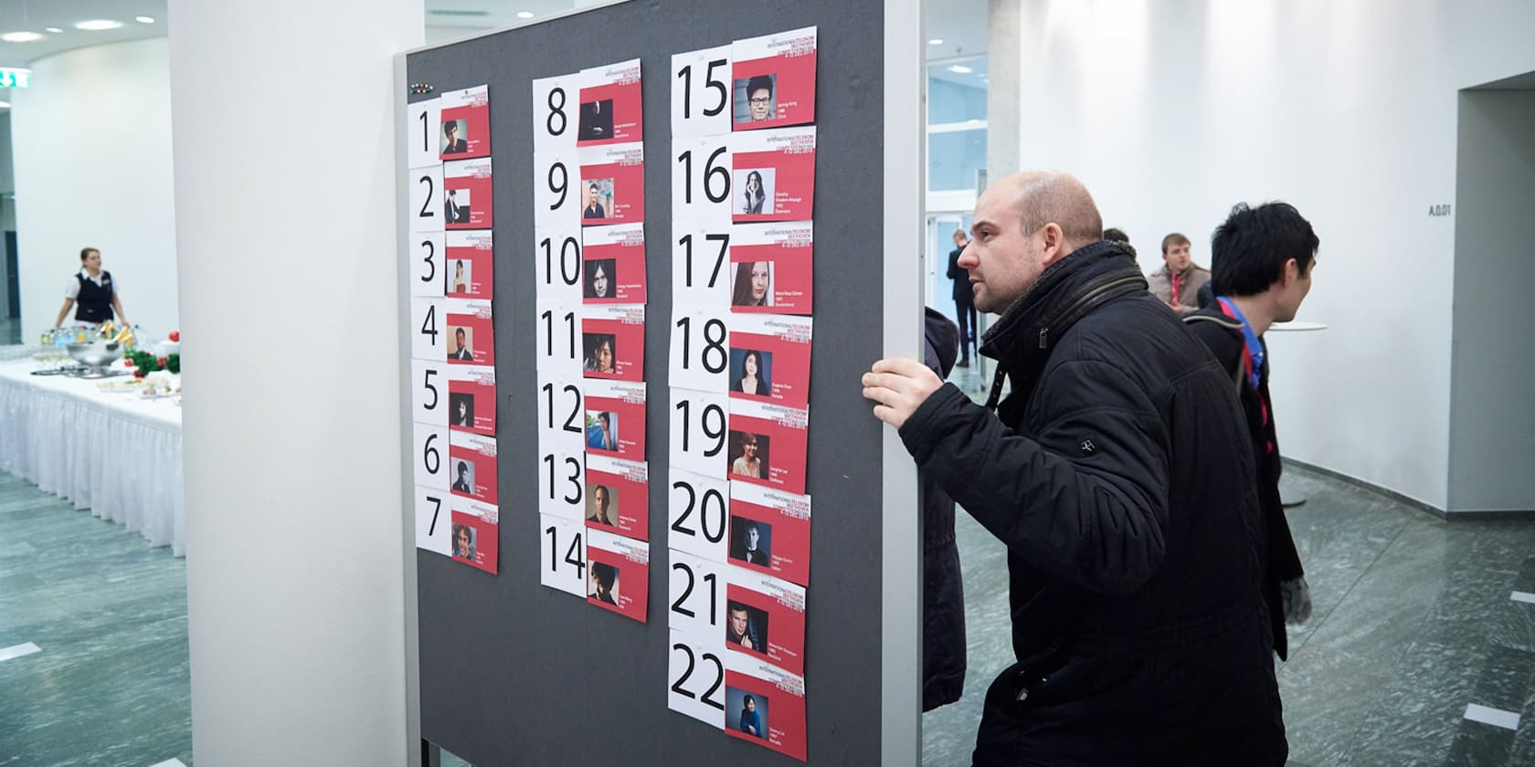 Mann betrachtet eine Tafel mit nummerierten Fotos und Namen von Personen in einer Ausstellungshalle.