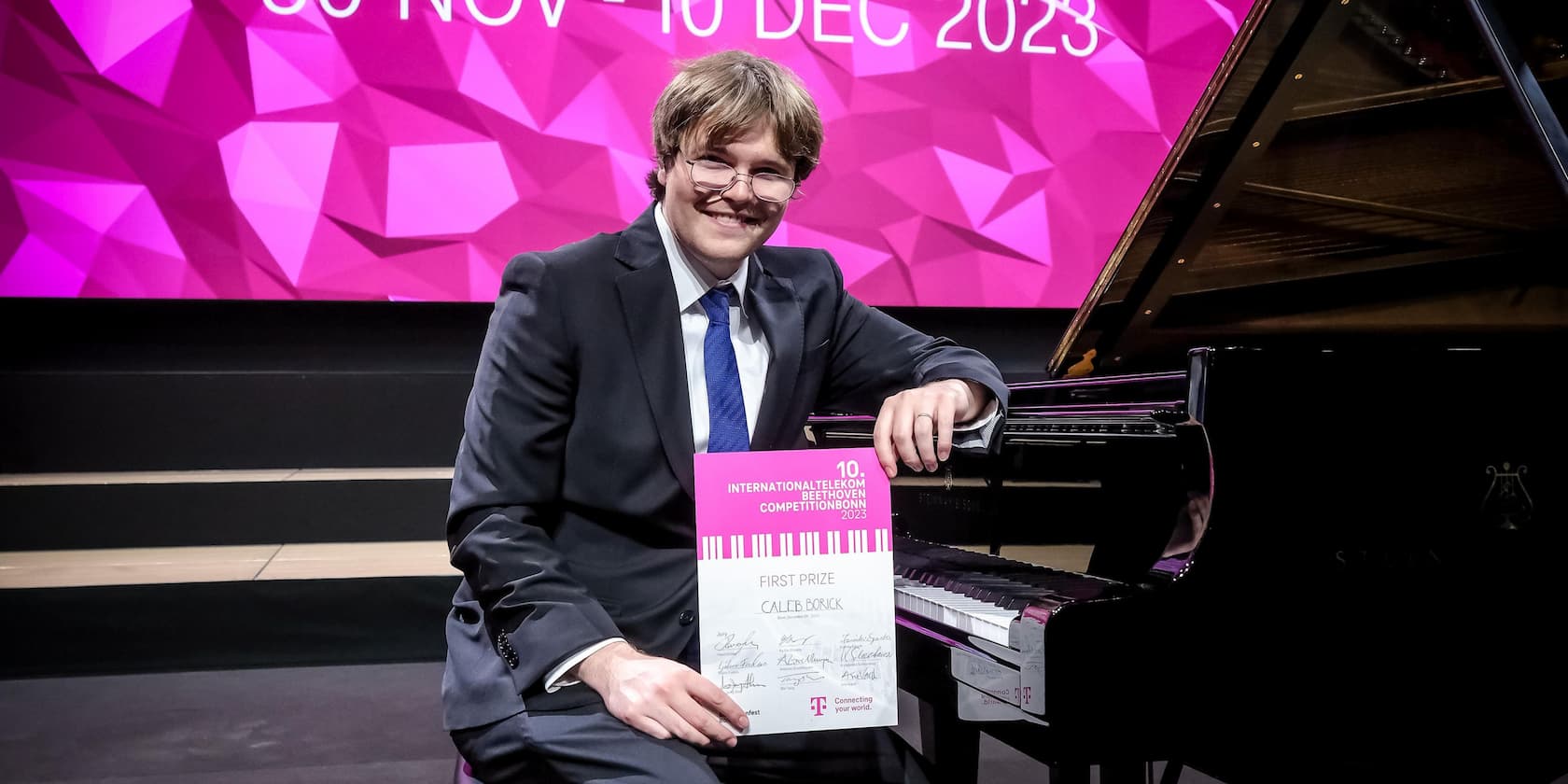 Ein Mann im Anzug sitzt vor einem Klavier und hält ein Zertifikat für den ersten Preis beim 10. Internationalen Telekom Beethoven Wettbewerb Bonn 2023.