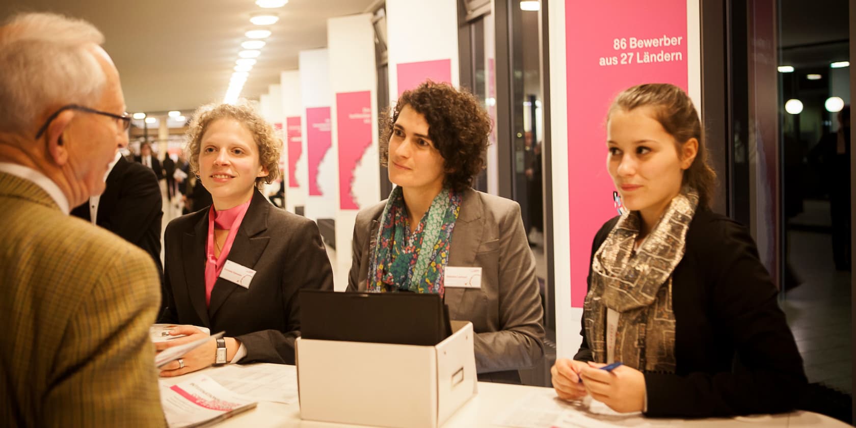 Three women at an information booth at a conference talking to an older man. In the background, a poster reads: '86 applicants from 27 countries'.