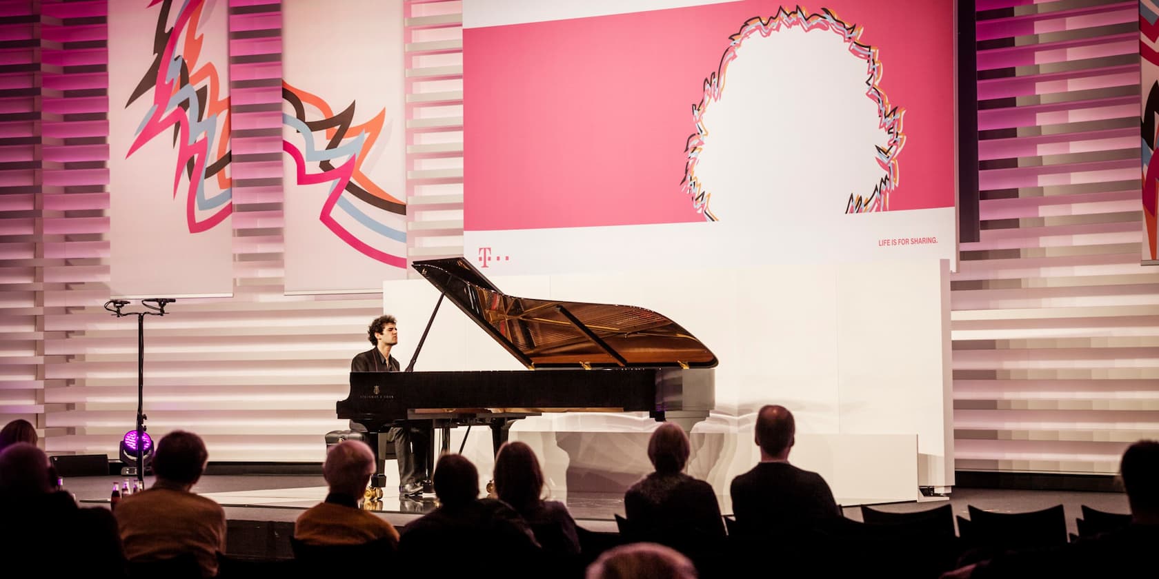 A musician plays piano on a stage during a presentation. In the background, there is a large, abstract artwork and the Telekom logo with the slogan 'Life is for sharing'. An audience is seated in front of the stage listening.