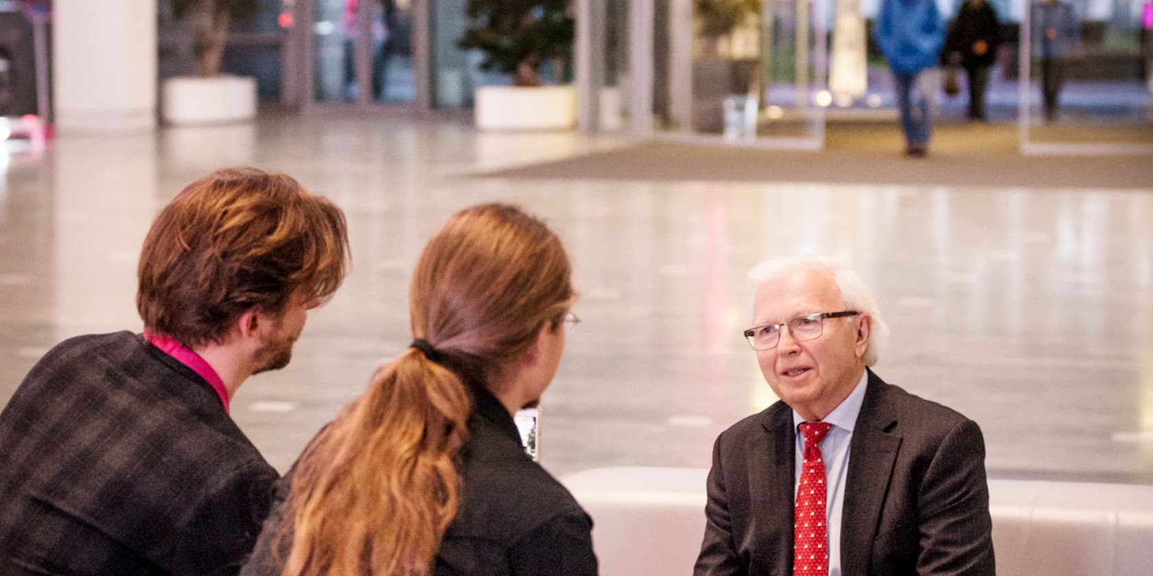 Drei Personen führen ein Gespräch in einer offenen Innenraumumgebung. Der ältere Mann in einem Anzug sitzt gegenüber den beiden anderen.