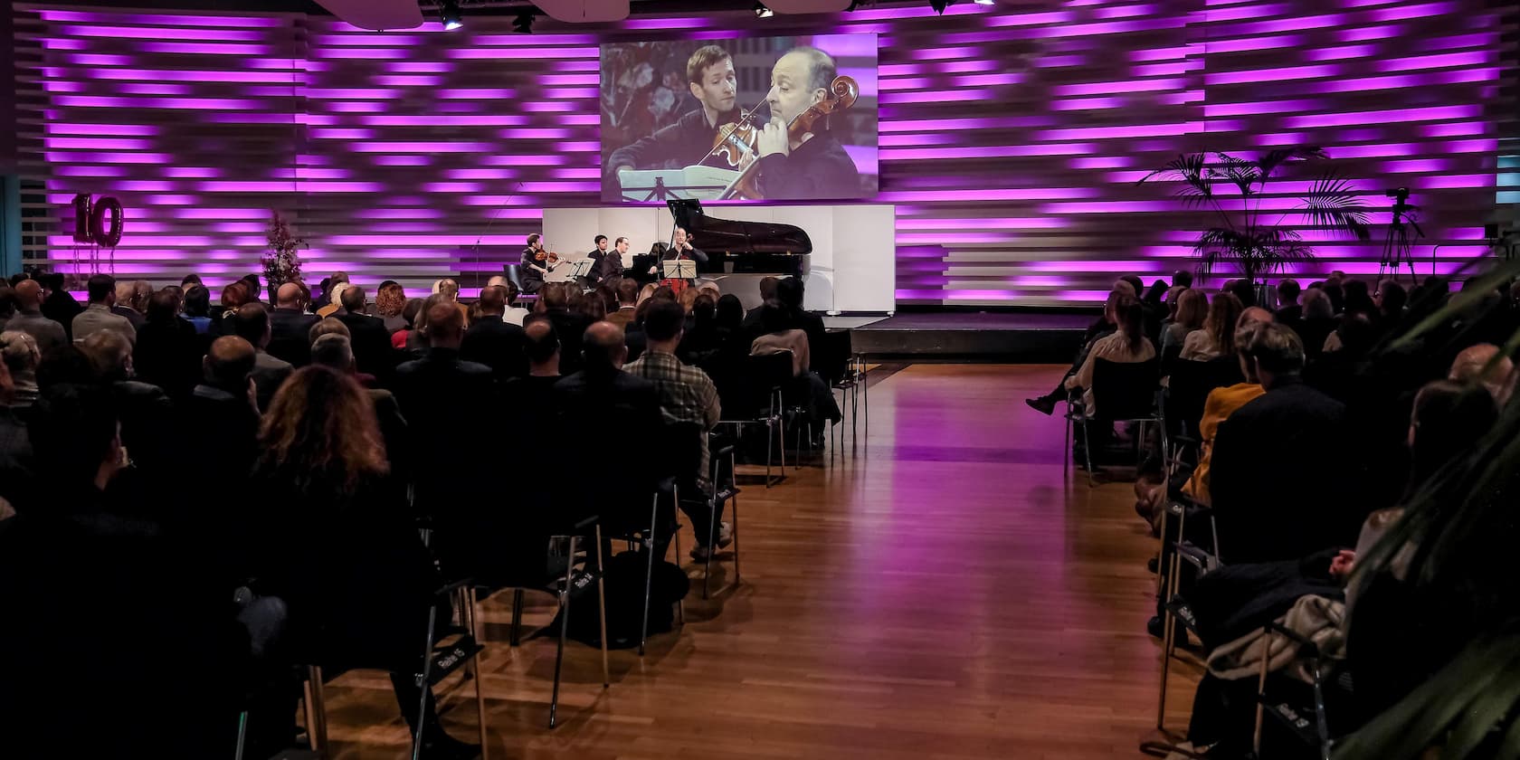 Ein Ensemble spielt Musik vor einem Publikum in einem hell erleuchteten Raum mit lila Beleuchtung, während im Hintergrund eine Projektion des Auftritts angezeigt wird.