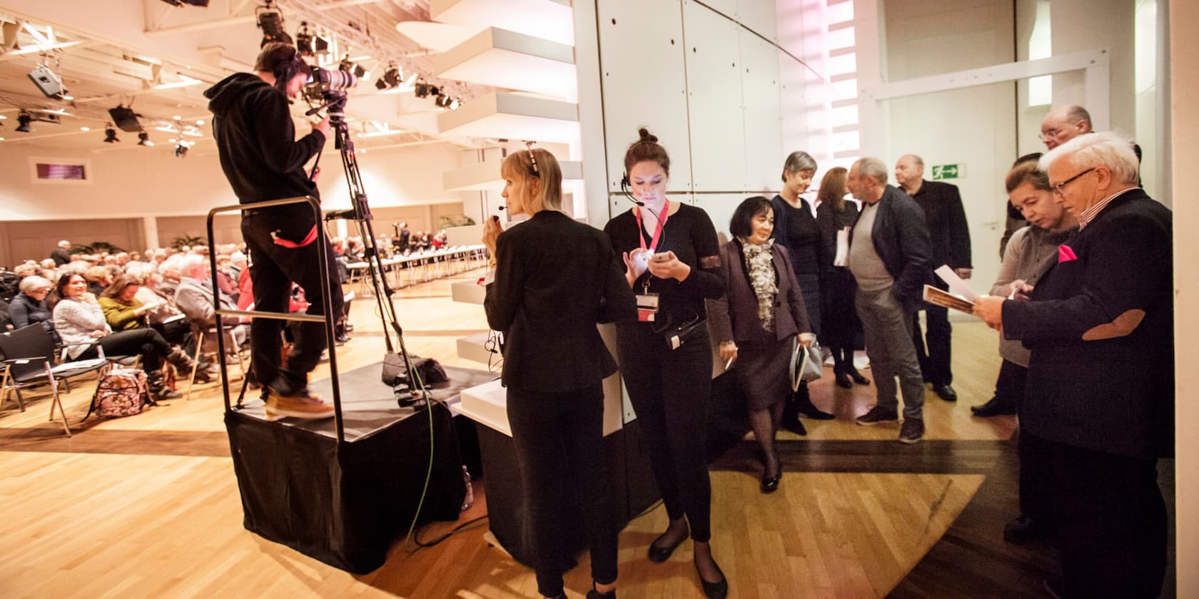 An event hall with seated audience, a cameraman, and several people conversing.