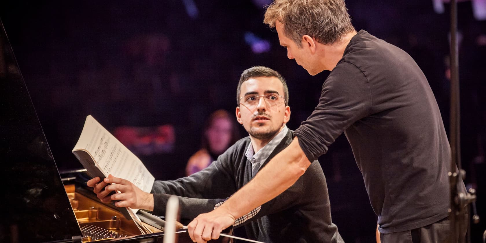 A man showing a sheet of music to another man at the piano.