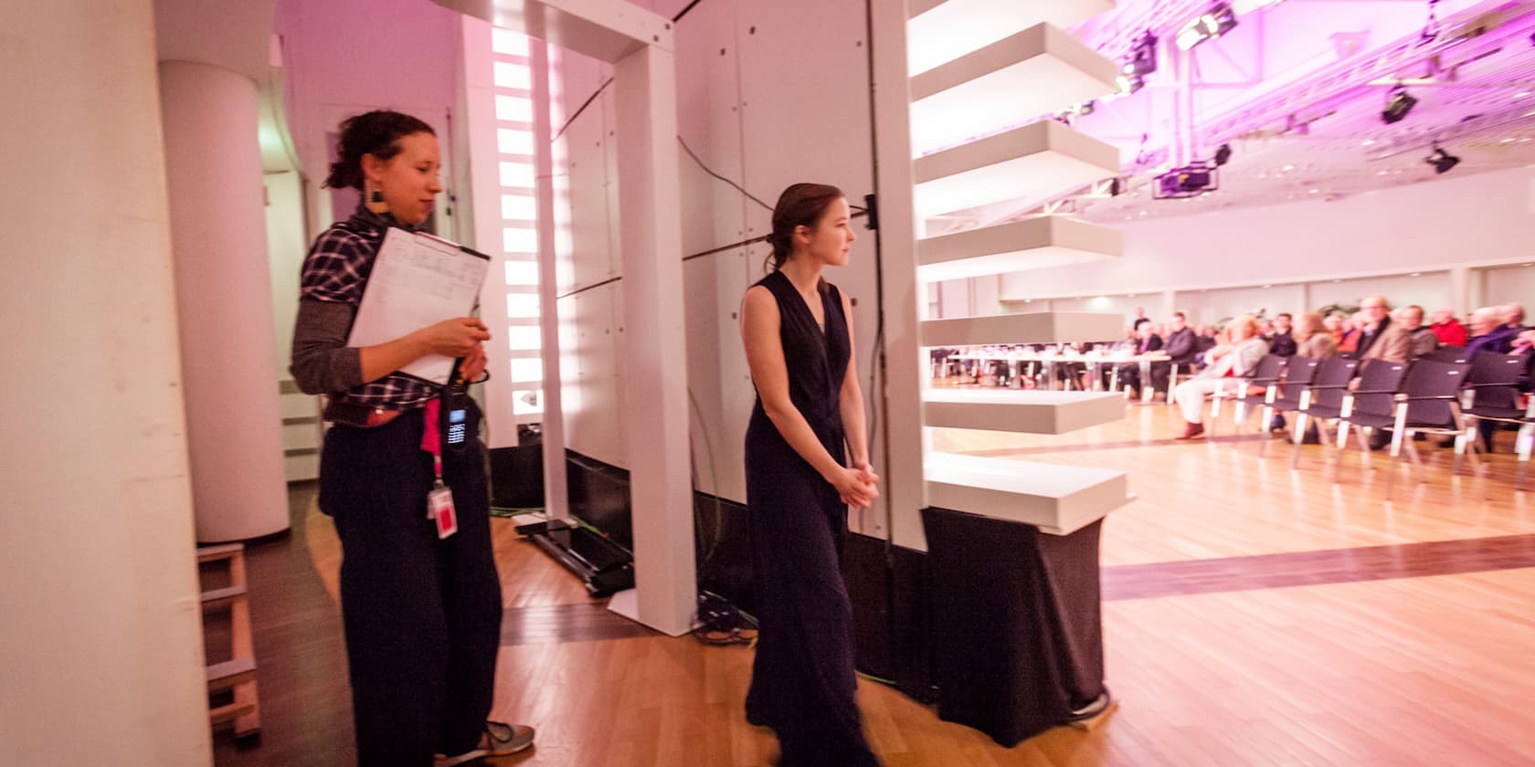 Two women stand backstage. One woman holds a clipboard, the other wears a long dress. Audience members are visible in the background.