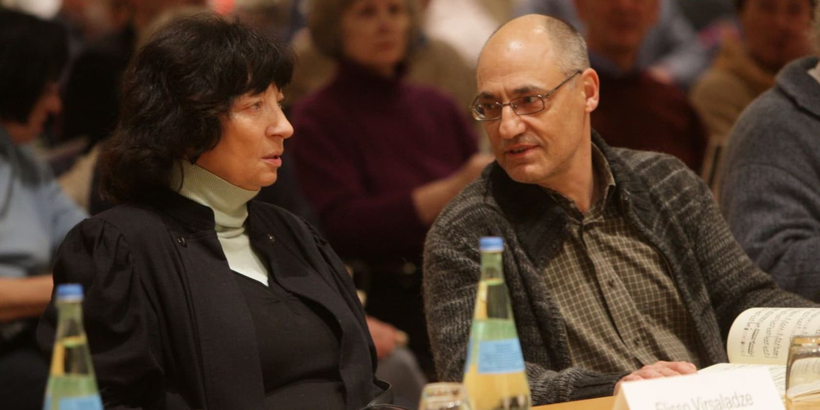 Two people sit at a table in an auditorium, talking to each other.