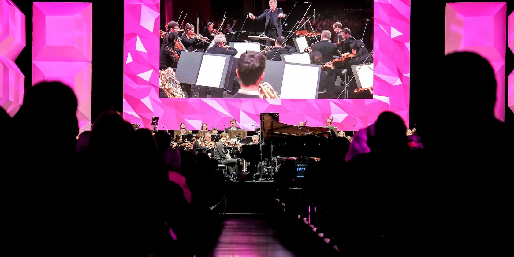 Orchestra performing on stage in front of an audience, with a large screen in the background