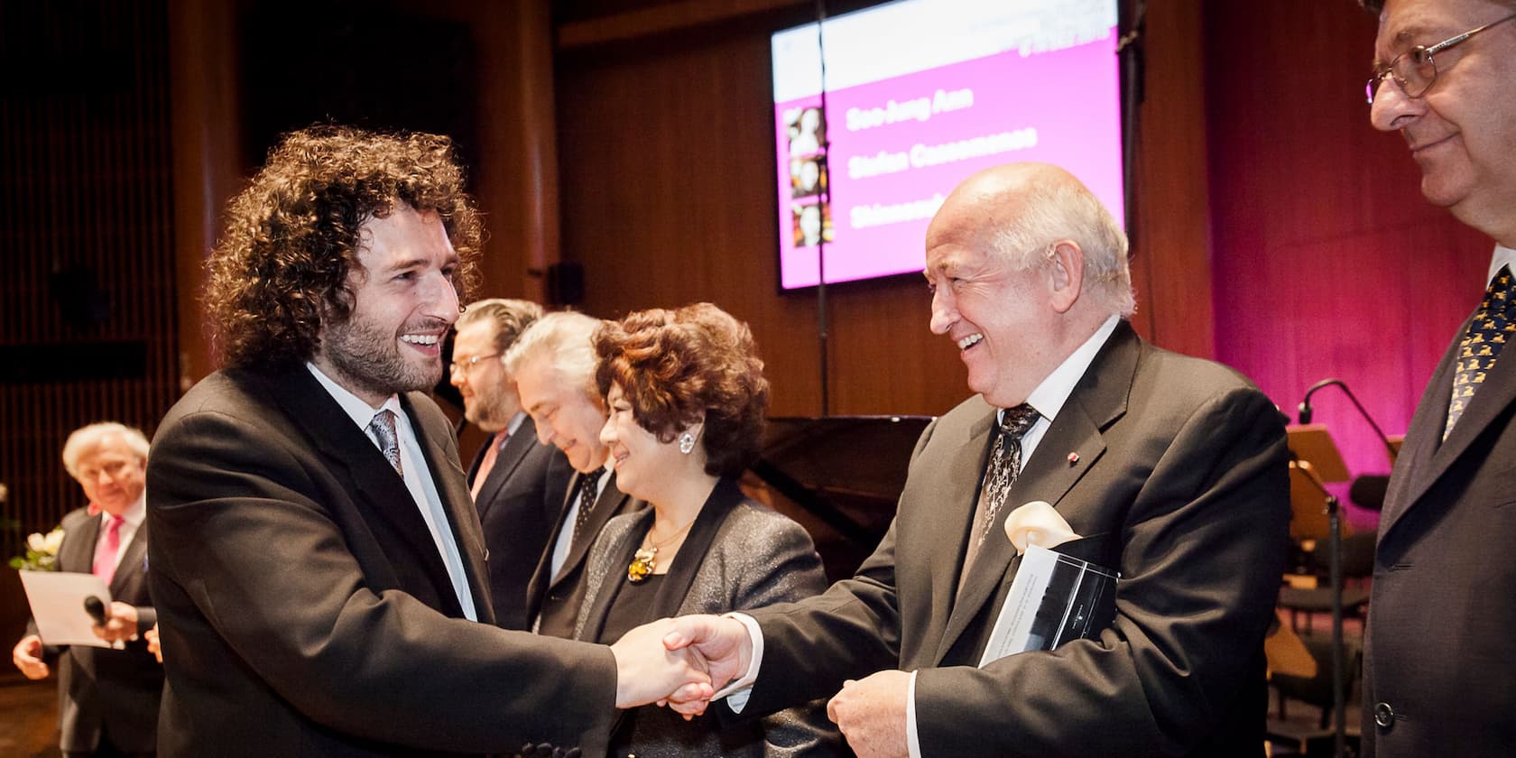 People in suits and formal attire shaking hands at an award ceremony.