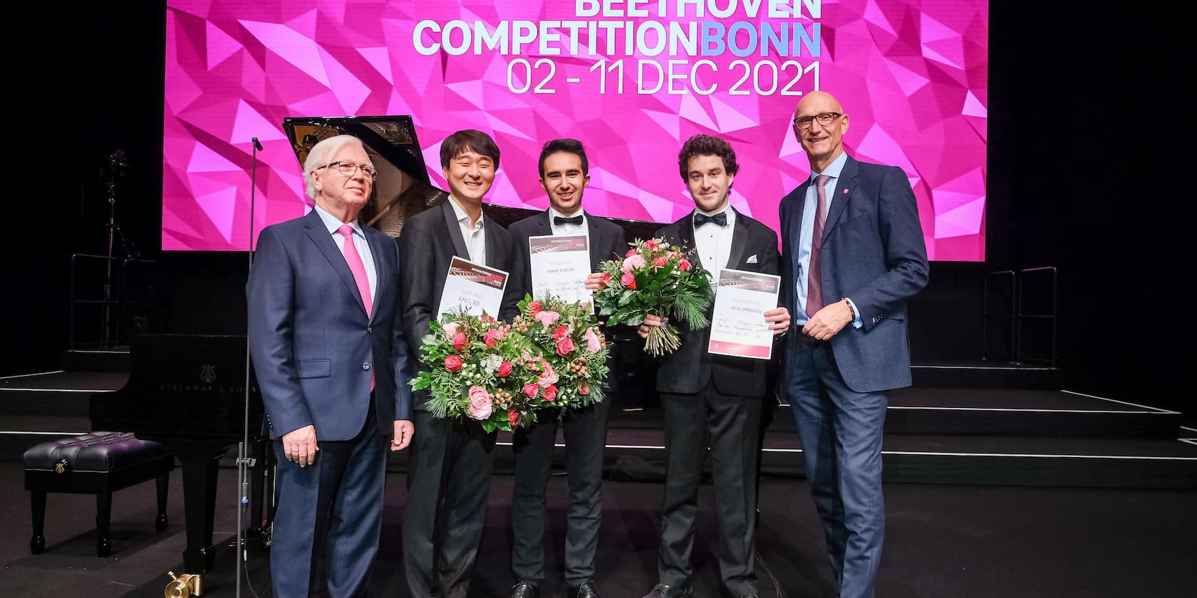 Award ceremony of the Beethoven Competition Bonn on December 11, 2021. Five men are standing on stage. Three of the men are holding bouquets and certificates.