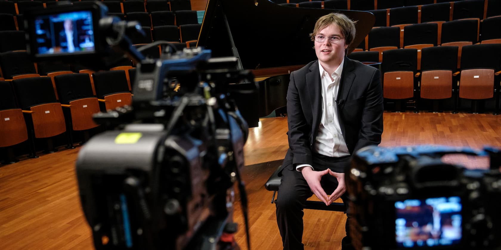 A man in a suit sits on a stage giving an interview in front of cameras.