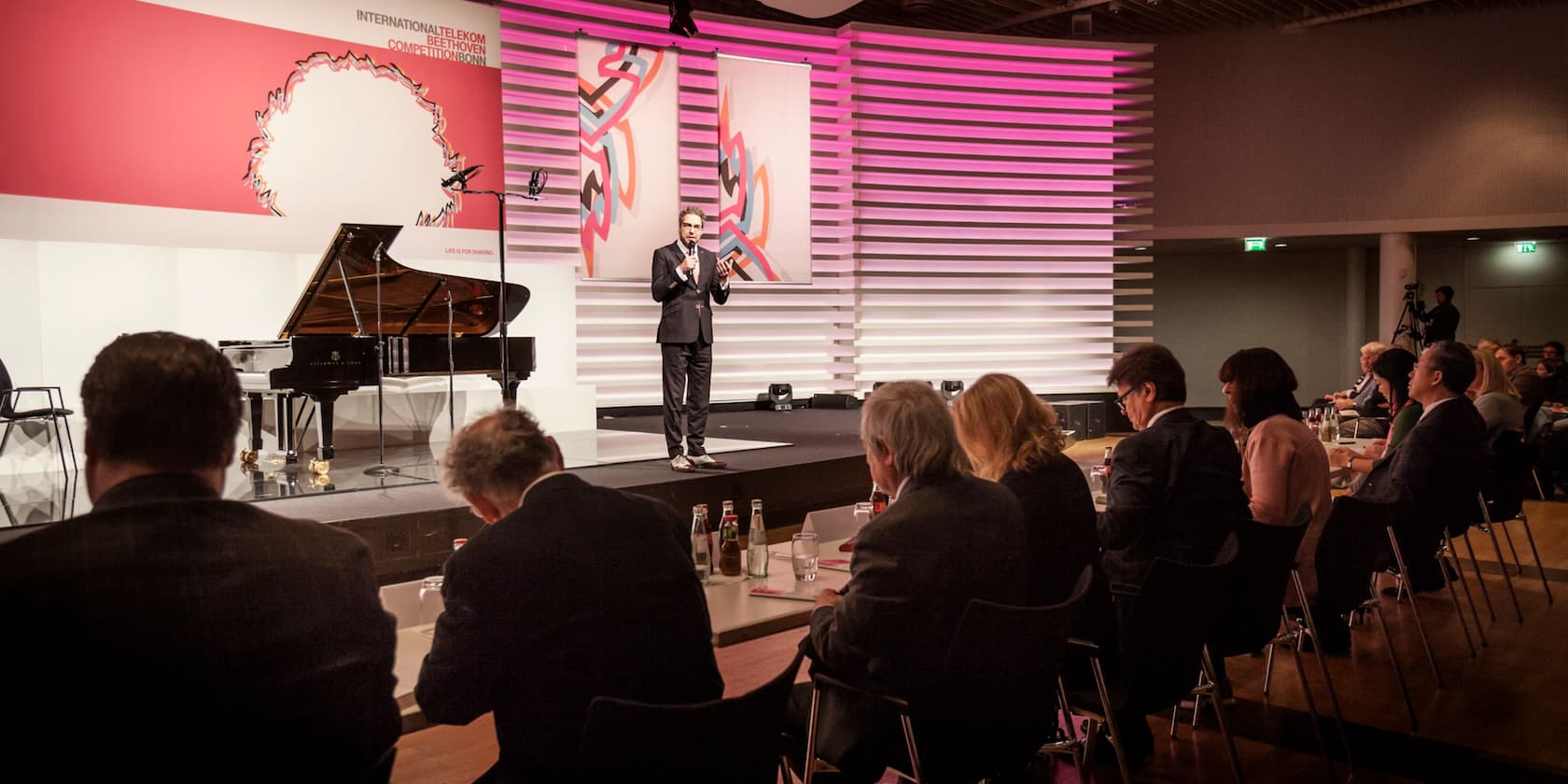 A man gives a speech on the stage of the International Telekom Beethoven Competition in Bonn. Audience members are seated at tables in the foreground.