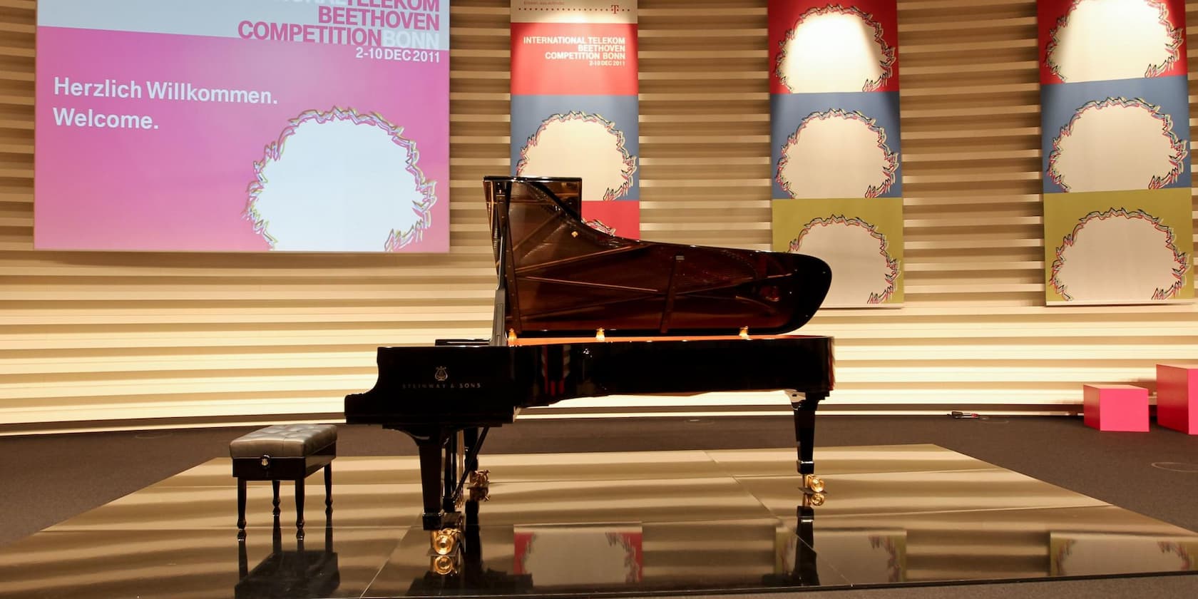 A grand piano is on a stage in front of a screen that reads 'Herzlich Willkommen. Welcome.' and the title 'International Telekom Beethoven Competition Bonn, 2-10 Dec 2011'.