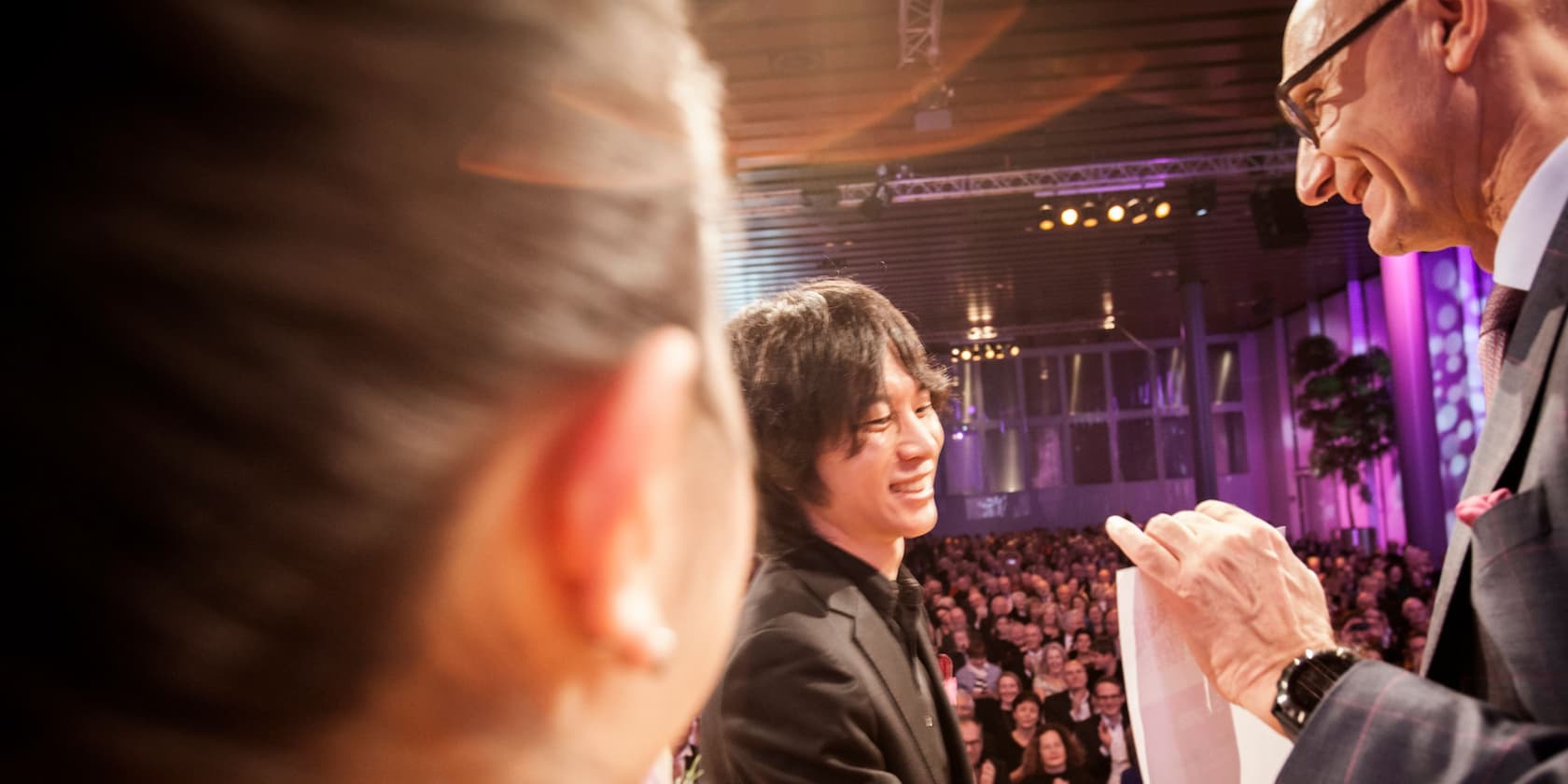 A man in a suit hands a document to another man at an event. Audience in the background.