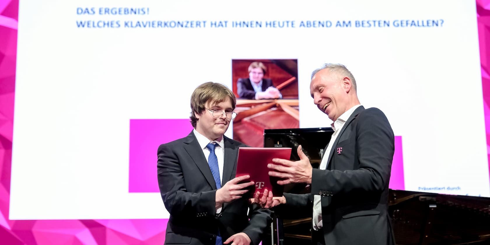 Two men in suits holding a red book in front of a screen with the text 'THE RESULT! WHICH PIANO CONCERT DID YOU ENJOY THE MOST THIS EVENING?'.