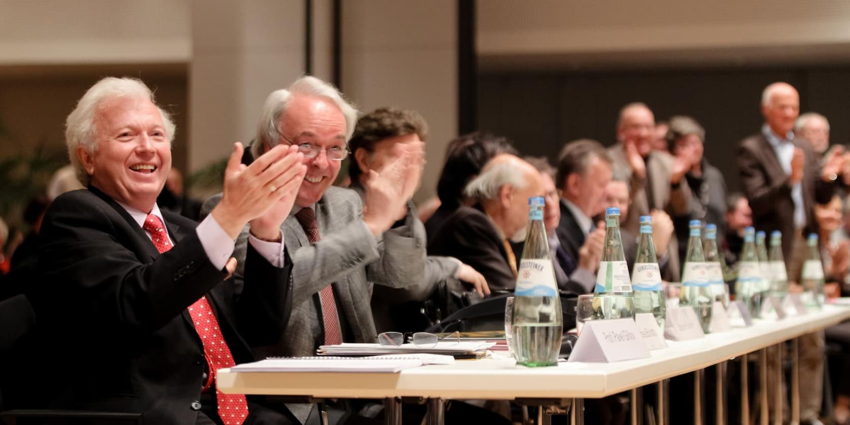 Men in suits sitting at a table and applauding at an event.