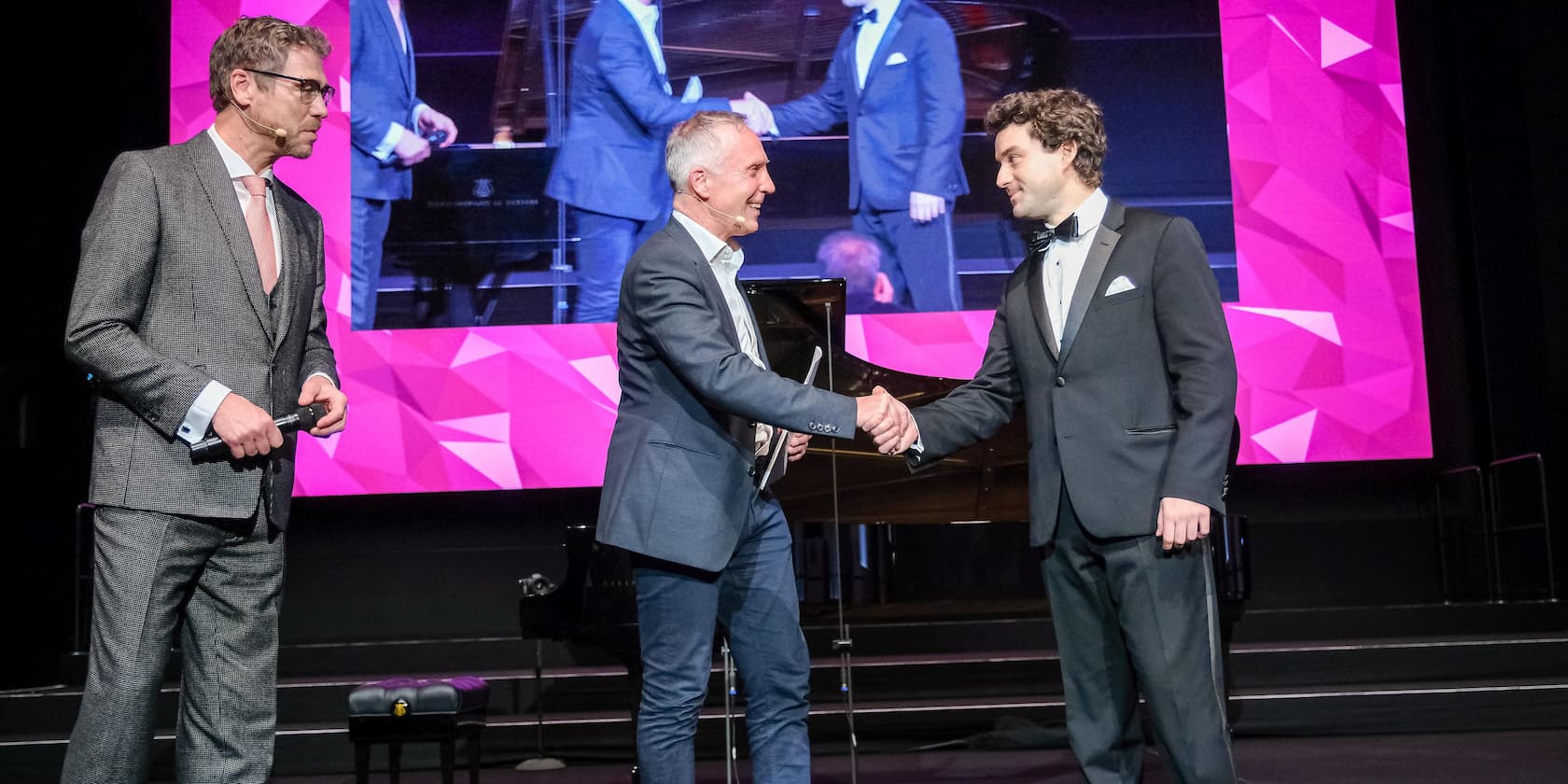 Three men on a stage at an awards ceremony, two of them shaking hands