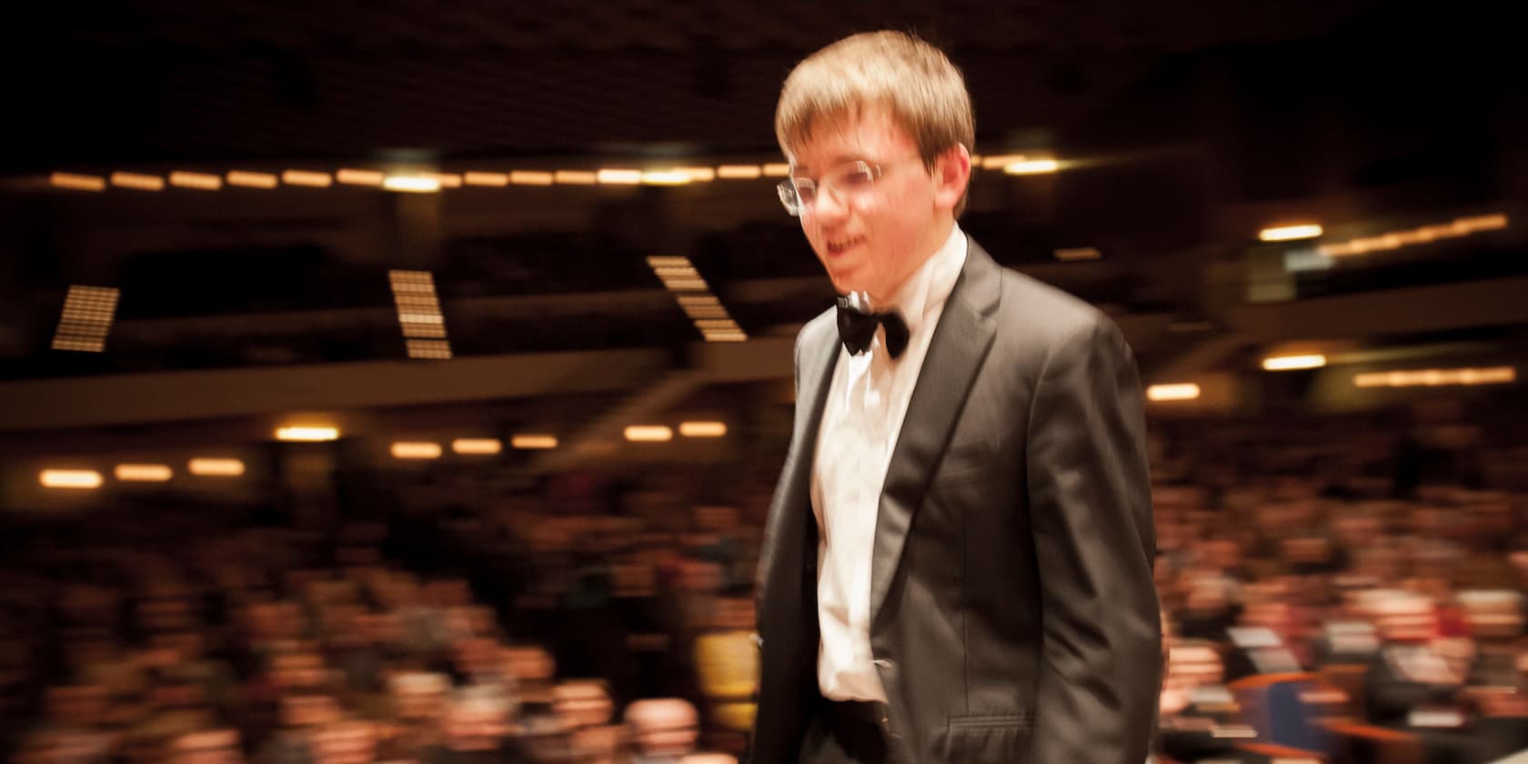 A man in a suit with a bow tie stands in front of a blurred audience in a large hall.