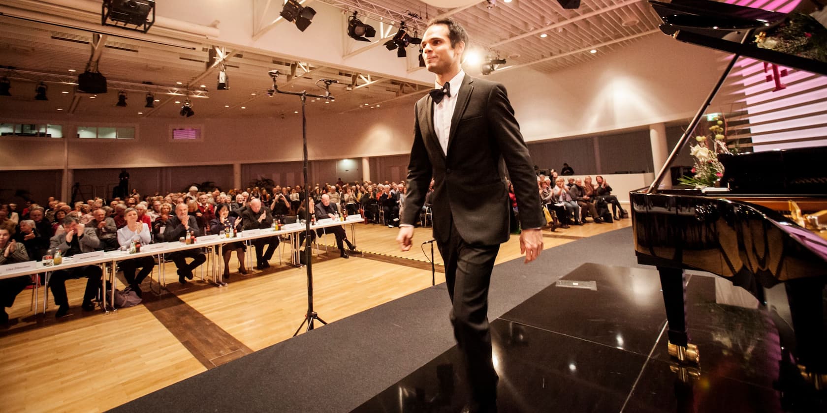 A man in a suit walks on a stage past a piano as the audience applauds.