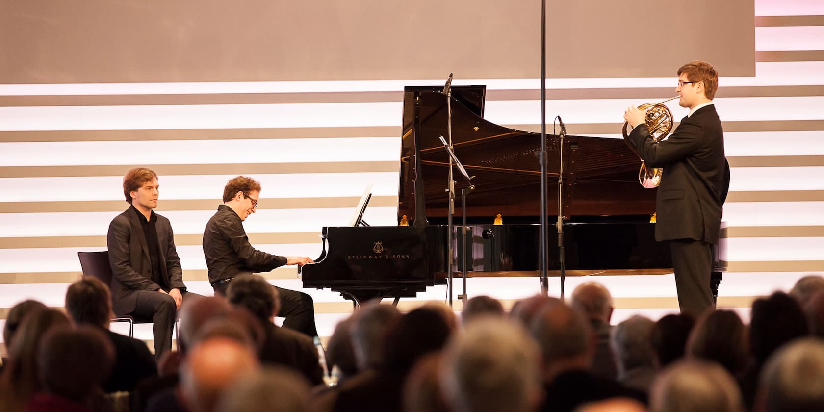 A pianist and a horn player perform in front of an audience. Another man sits on a chair next to the pianist.