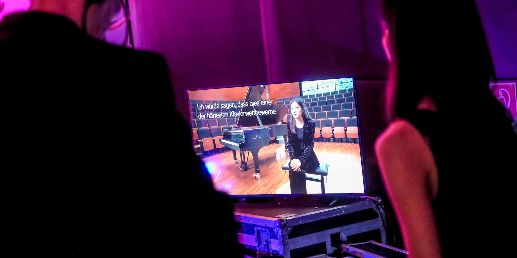 Two people are watching a woman on a screen talking about piano competitions. The text on the screen reads: 'Ich würde sagen, dass dies einer der härtesten Klavierwettbewerbe ist.'