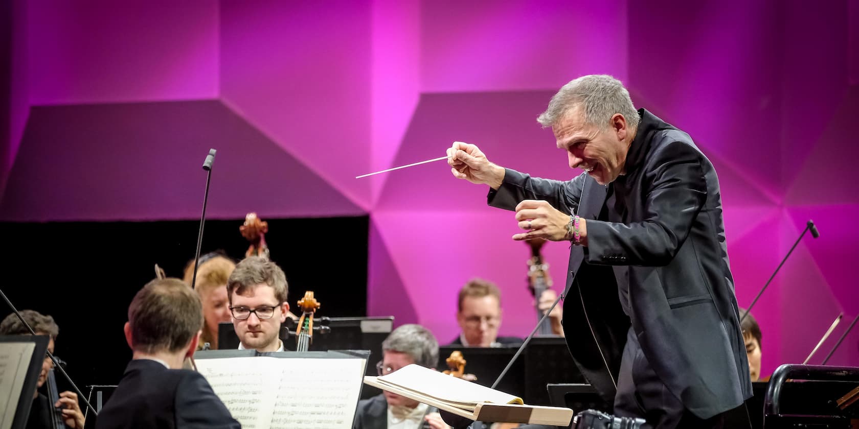 A conductor leads an orchestra on a stage with a purple background.