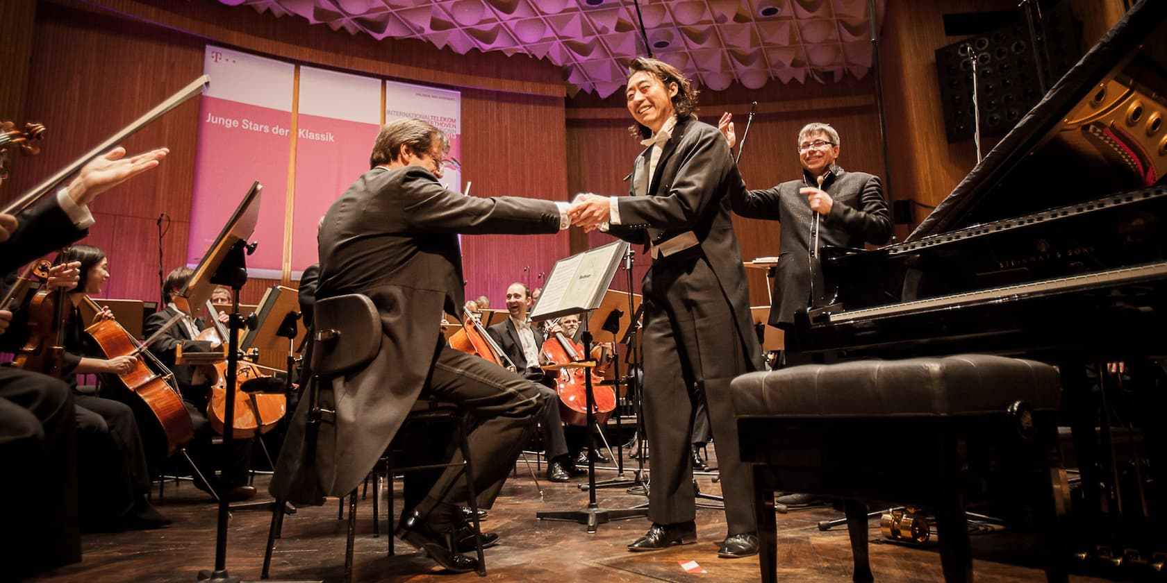 Musicians at a classical concert, two men shaking hands in front of the orchestra. In the background, a banner with the text 'Junge Stars der Klassik' is visible.