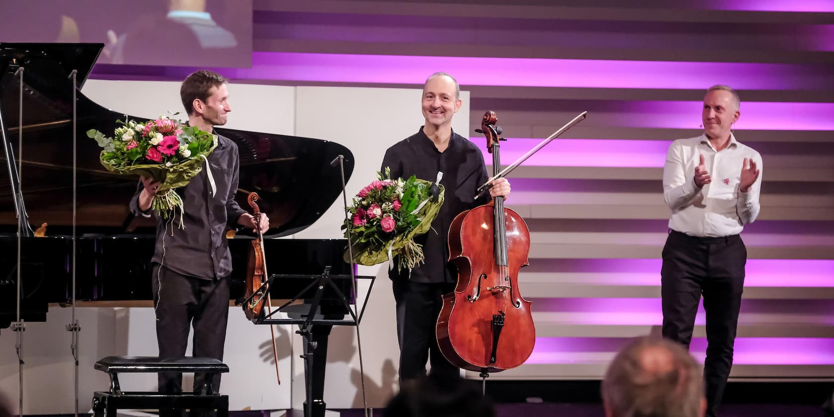 Three men on a stage. Two musicians hold bouquets and musical instruments, while a third man applauds.