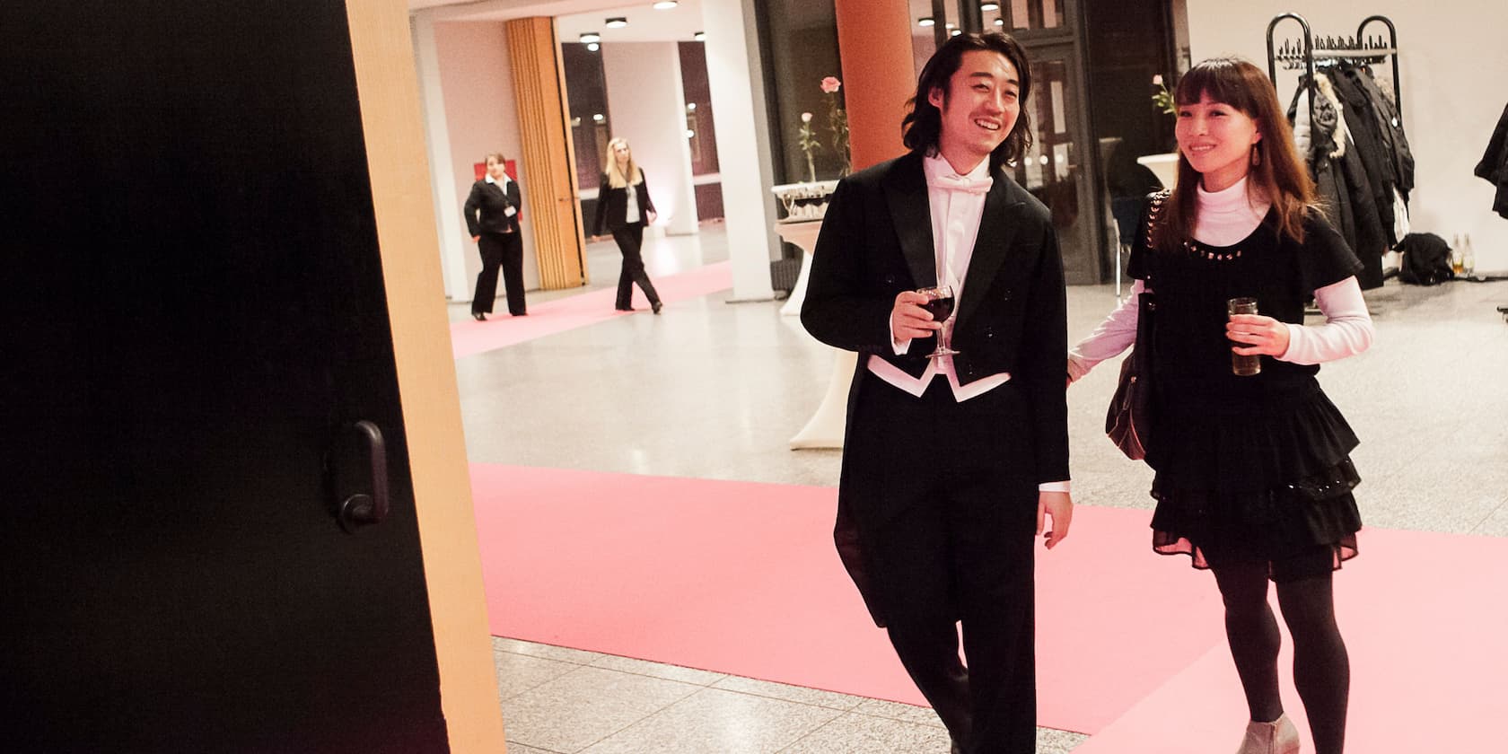 A man in a tuxedo and a woman in a black dress walk on a red carpet at an elegant event.