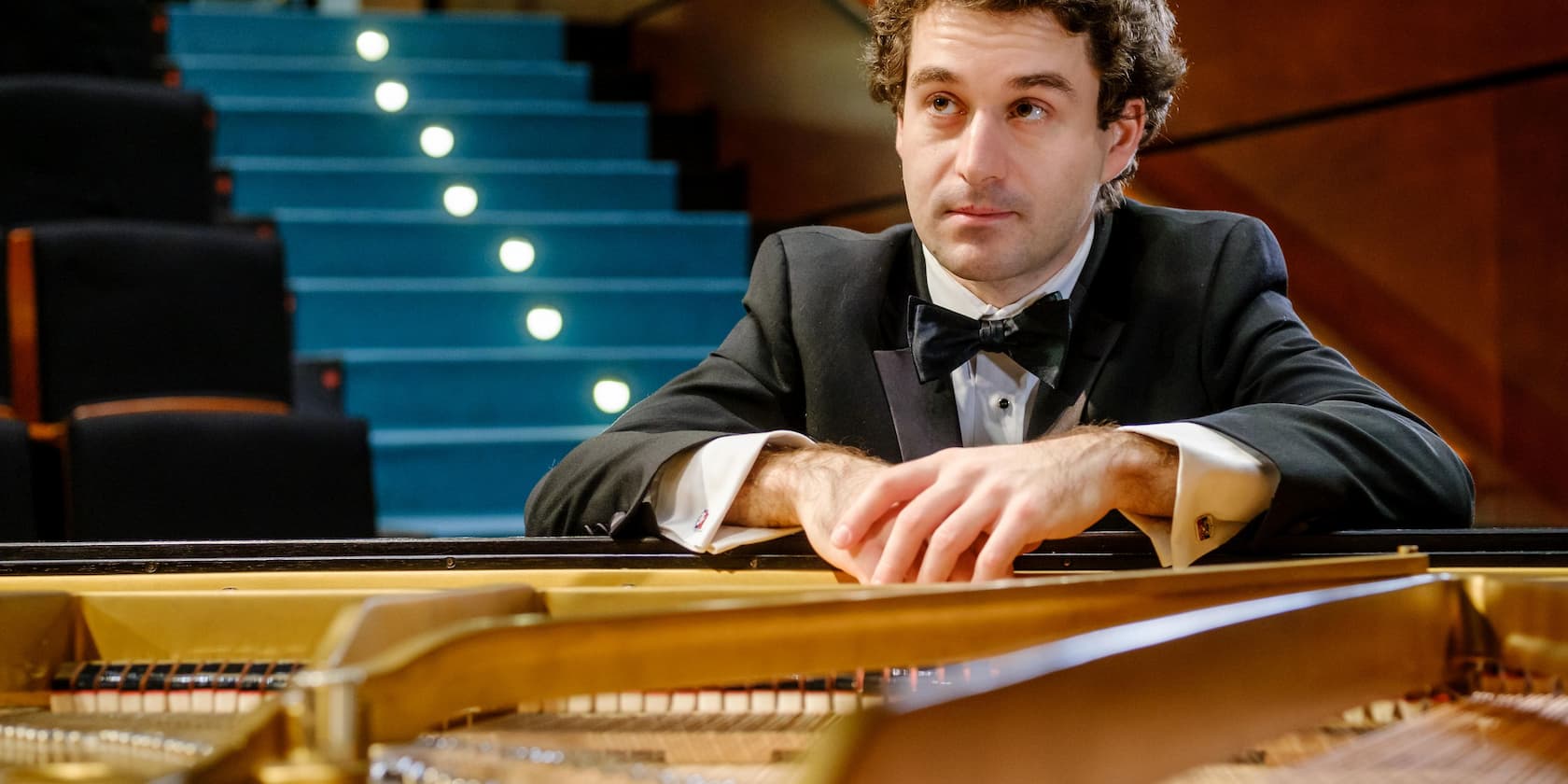 A man in a tuxedo sits at a piano, with a blue staircase in the background.