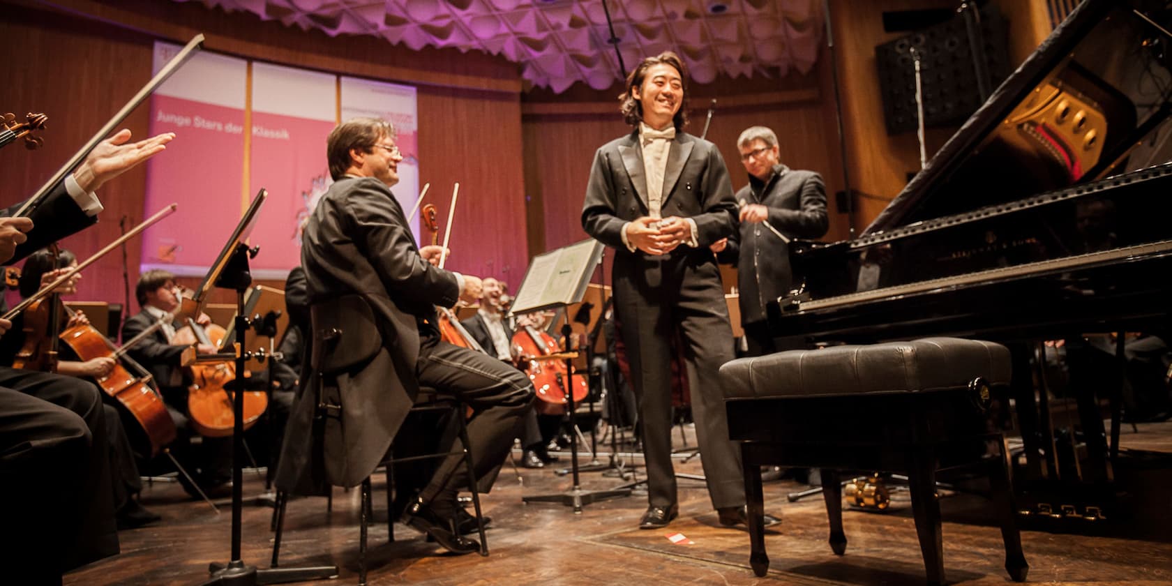 Orchestra playing on stage, conductor standing smiling to the right of the piano. In the background, a sign reads 'Young Stars of Classical Music'.