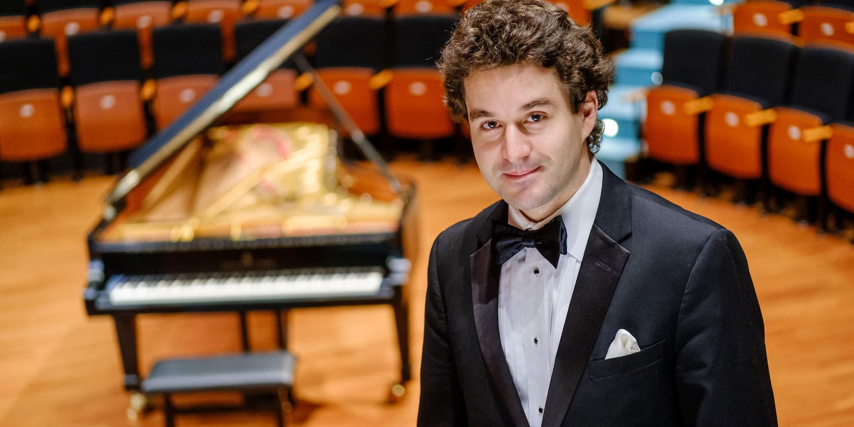 A man in a tuxedo stands on a stage in front of a grand piano, with empty theater seats in the background.