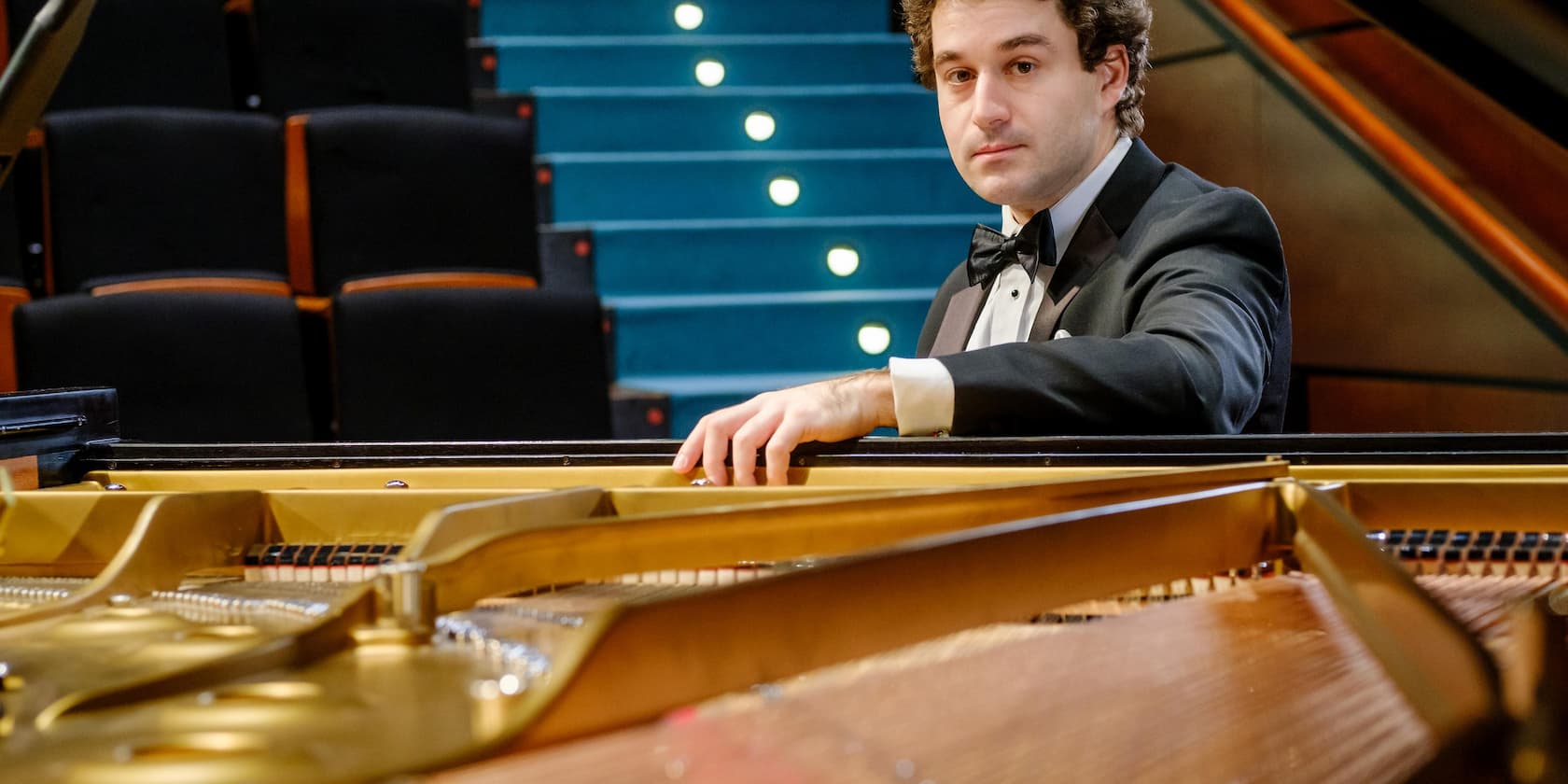 A pianist in a tuxedo plays a grand piano in front of an empty audience stand.