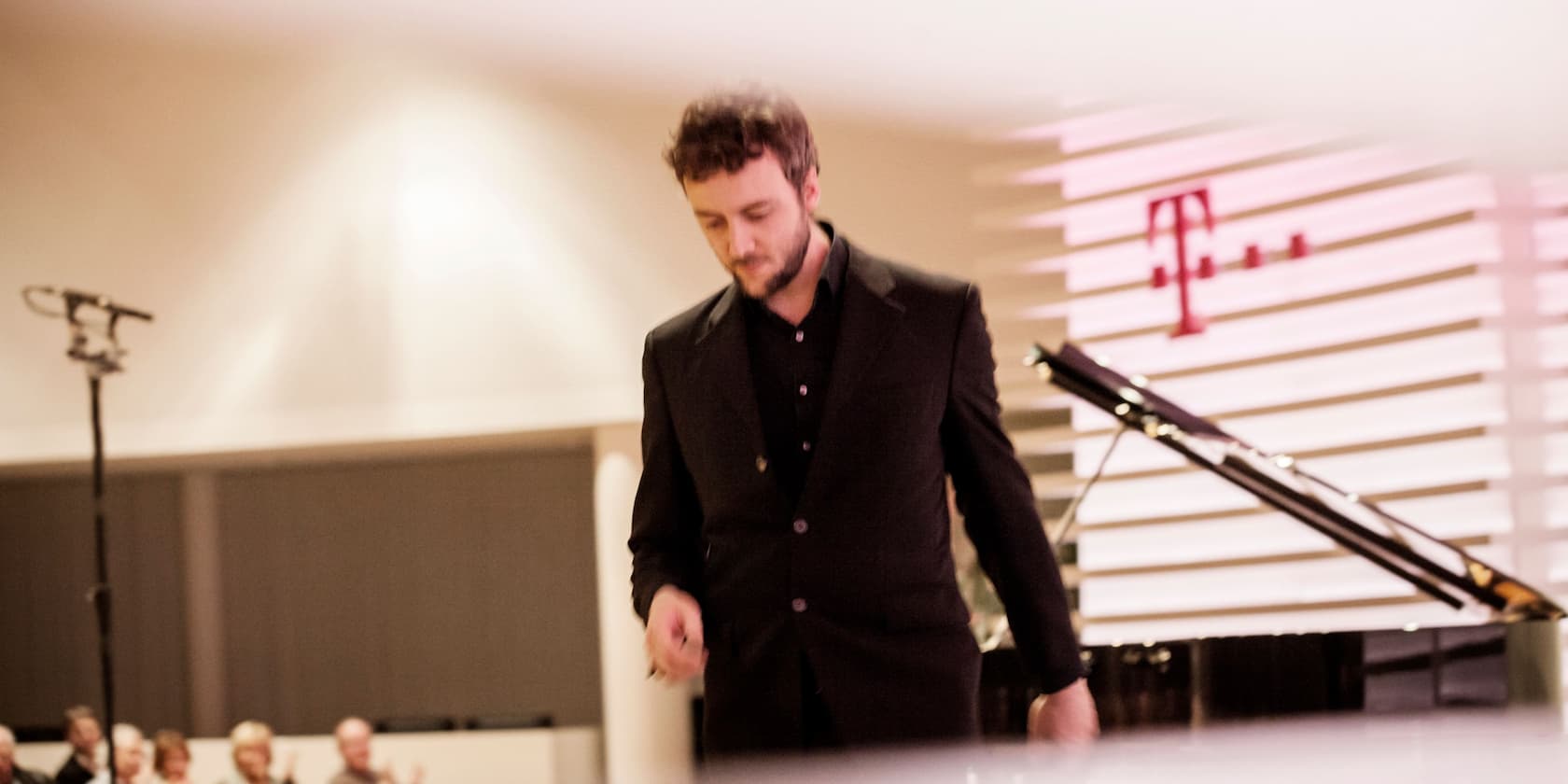 A man in a suit stands next to a grand piano. In the background, there is a microphone and an audience.