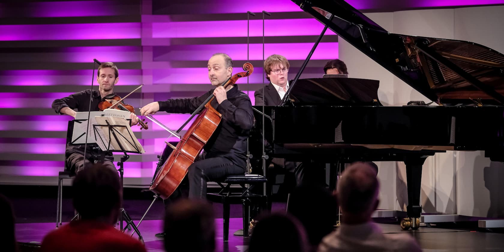 A trio of violin, cello, and piano performs on stage in front of an audience. The background is lit in shades of purple.