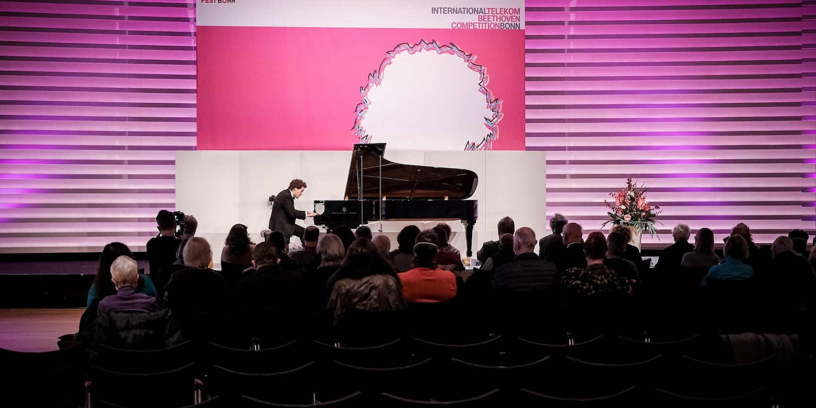 A pianist performs on stage during a competition in front of an audience. The background displays 'INTERNATIONAL TELEKOM BEETHOVEN COMPETITION BONN'.