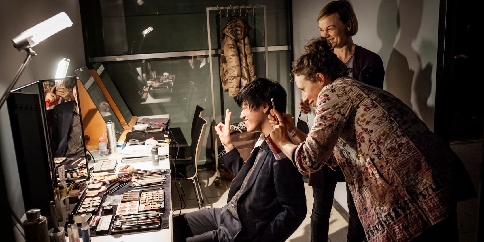 Two makeup artists are applying makeup to a man in front of a vanity table with makeup products.