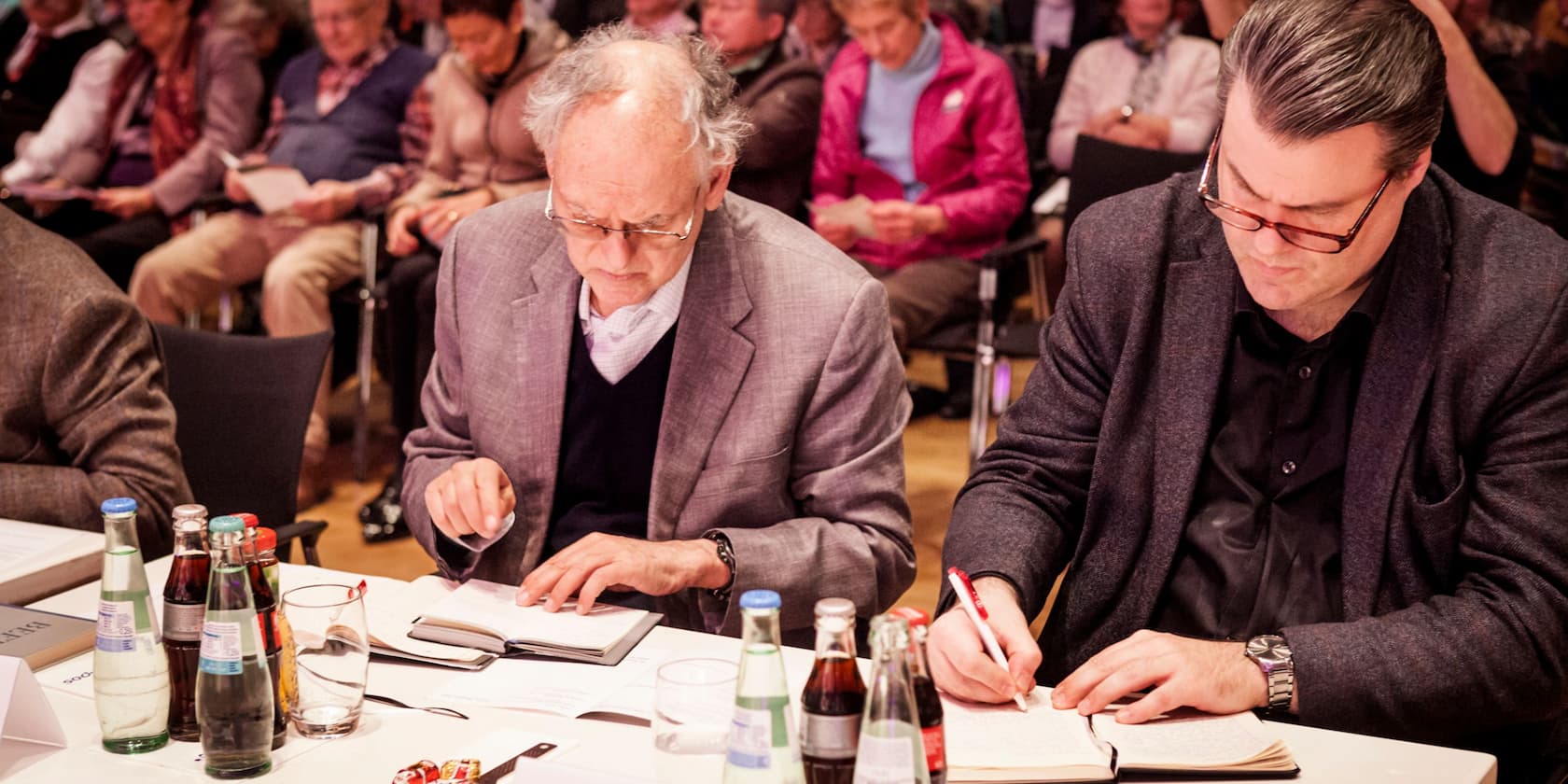 Two men are sitting at a table and writing in notebooks, with several people seated in the background.