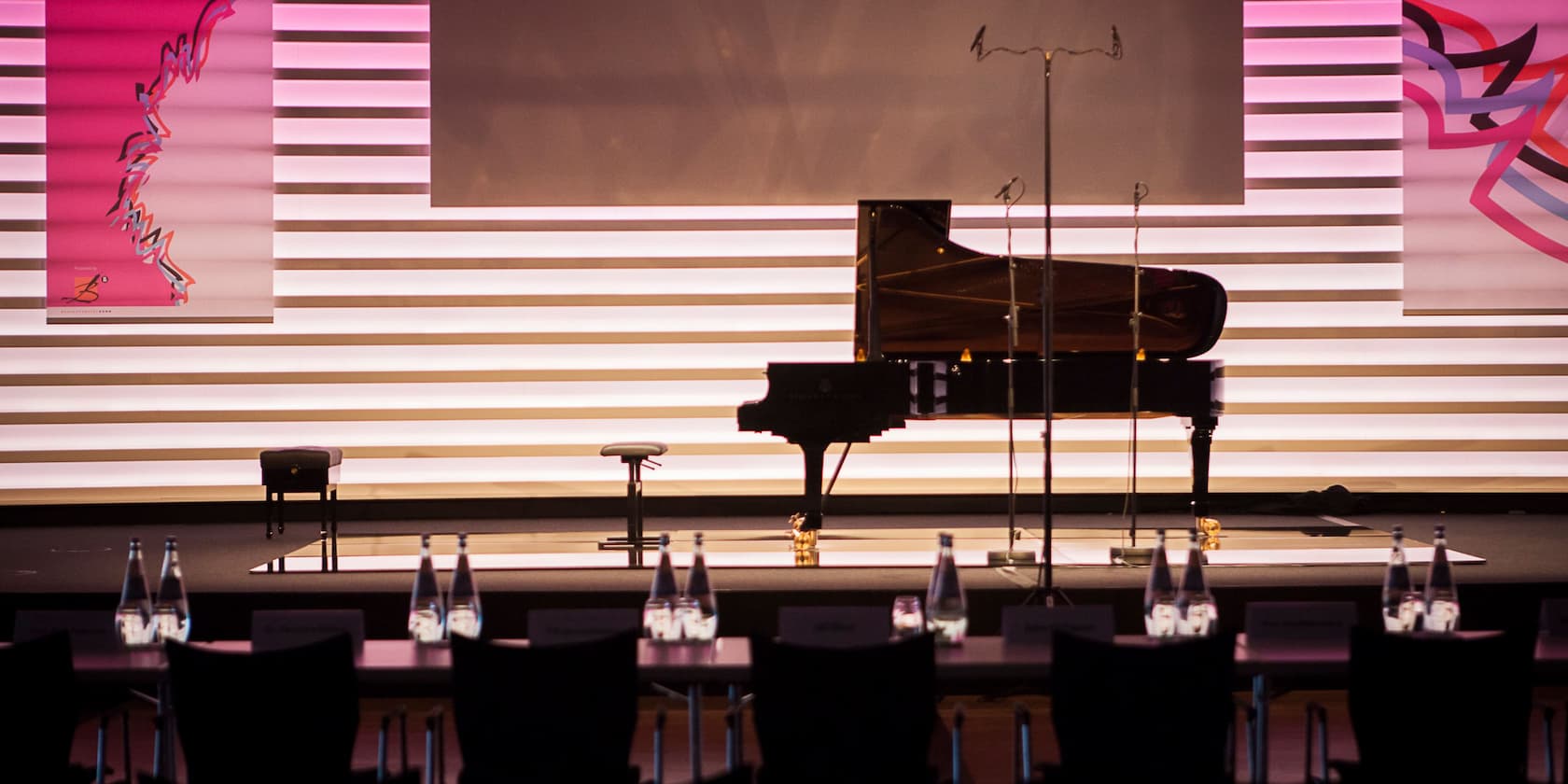 A concert hall with a grand piano on stage, surrounded by white and pink panels, with tables and water bottles in the foreground.